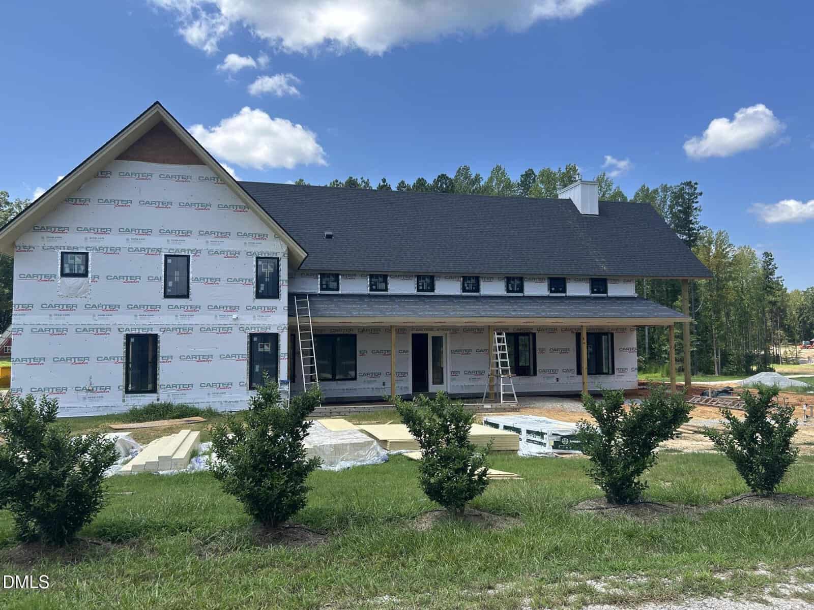 A large house under construction with scaffolding, ladders, and building materials scattered in the front yard. The house is partially wrapped in Carter paper, and shrubbery lines the foreground. Blue sky with clouds is visible.