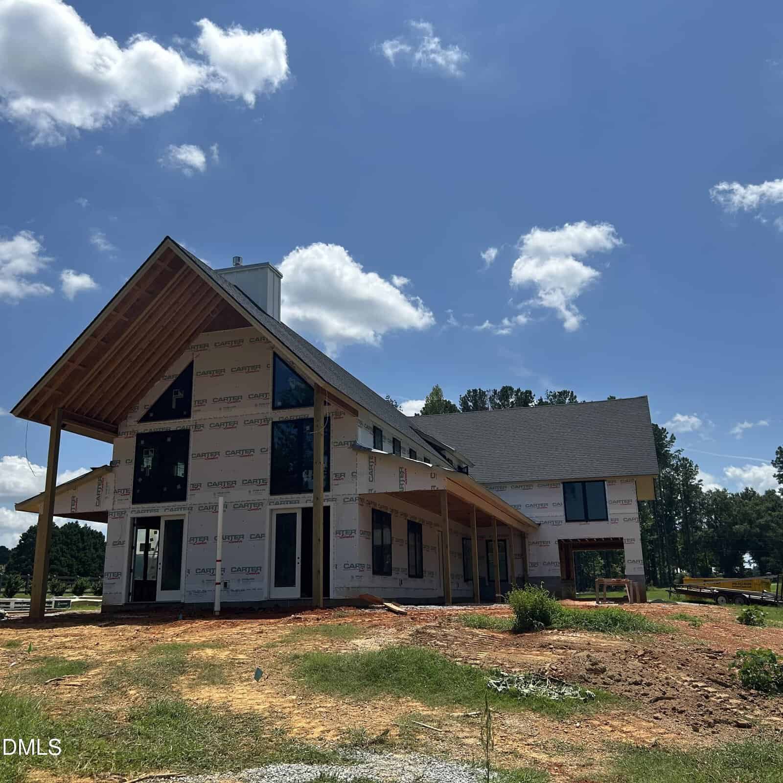 A two-story house under construction, partially covered with weatherproof sheeting and lacking exterior siding, sits on a dirt lot with some grass. The roof is completed, and windows have been installed. Trees are visible in the background.