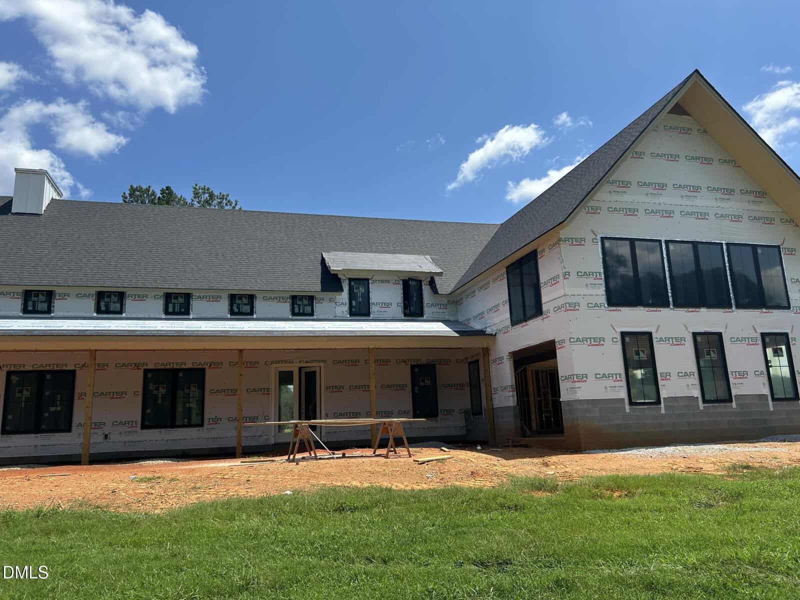 A large two-story house under construction, with house wrap covering the exterior walls, unfinished windows, and construction materials on the ground in front. The sky is clear with a few clouds.