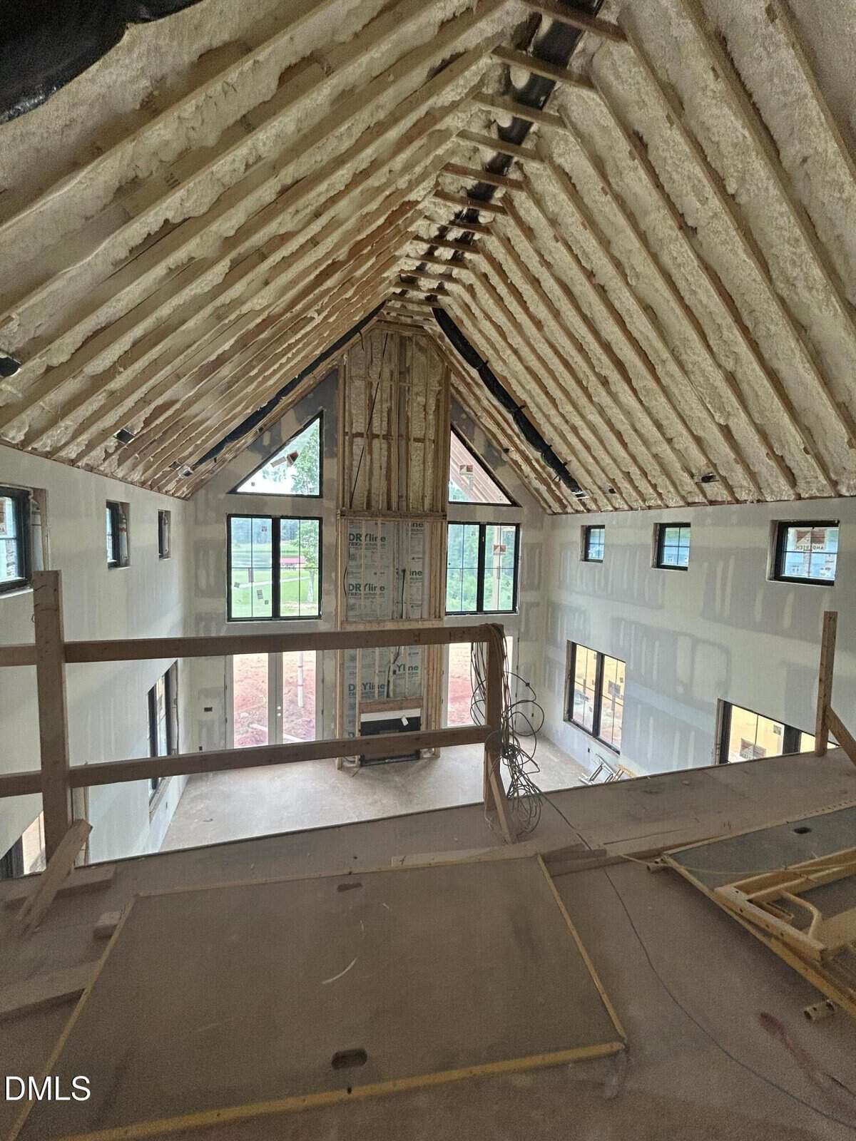 A view from a loft of a large house under construction, featuring exposed foam insulation in the vaulted ceiling, unfinished walls, and large windows letting in natural light.