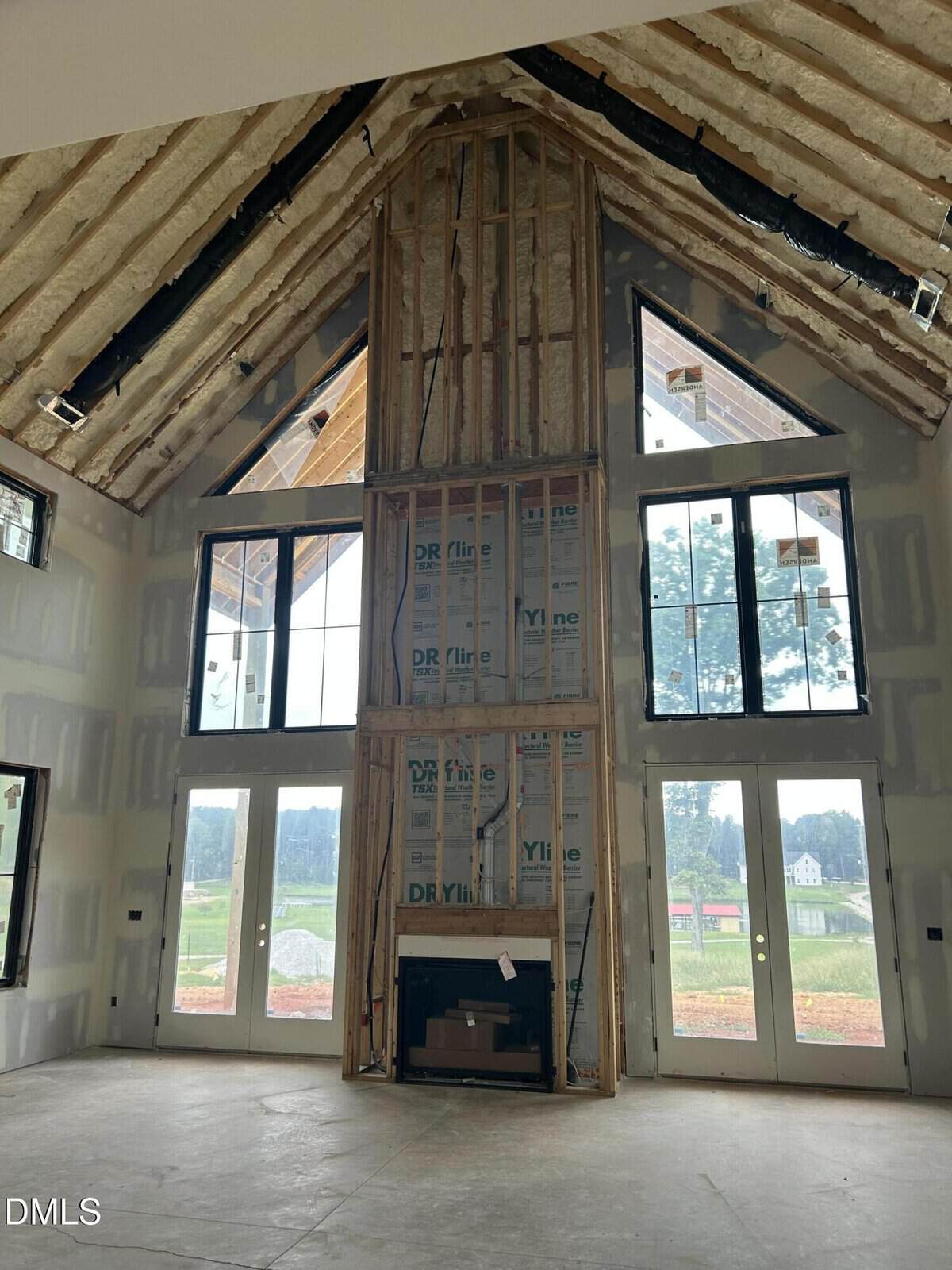 A tall, unfinished living room with exposed insulation, a central fireplace framed by large windows, and double glass doors overlooking a green outdoor area.