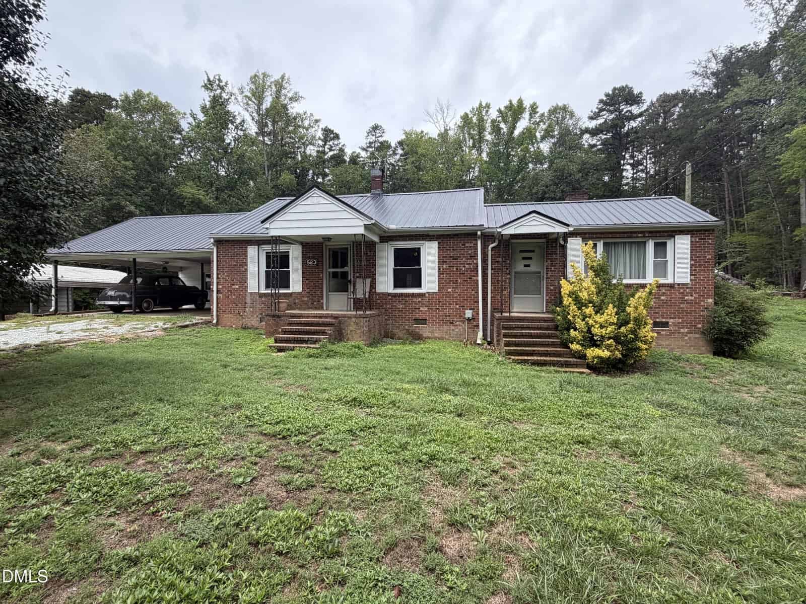 A single-story brick house with a metal roof, front porch steps, and a covered carport on the left sits at 523 US Highway 158 in Rougemont. The yard is grassy with some patches, and trees line the background under an overcast sky.