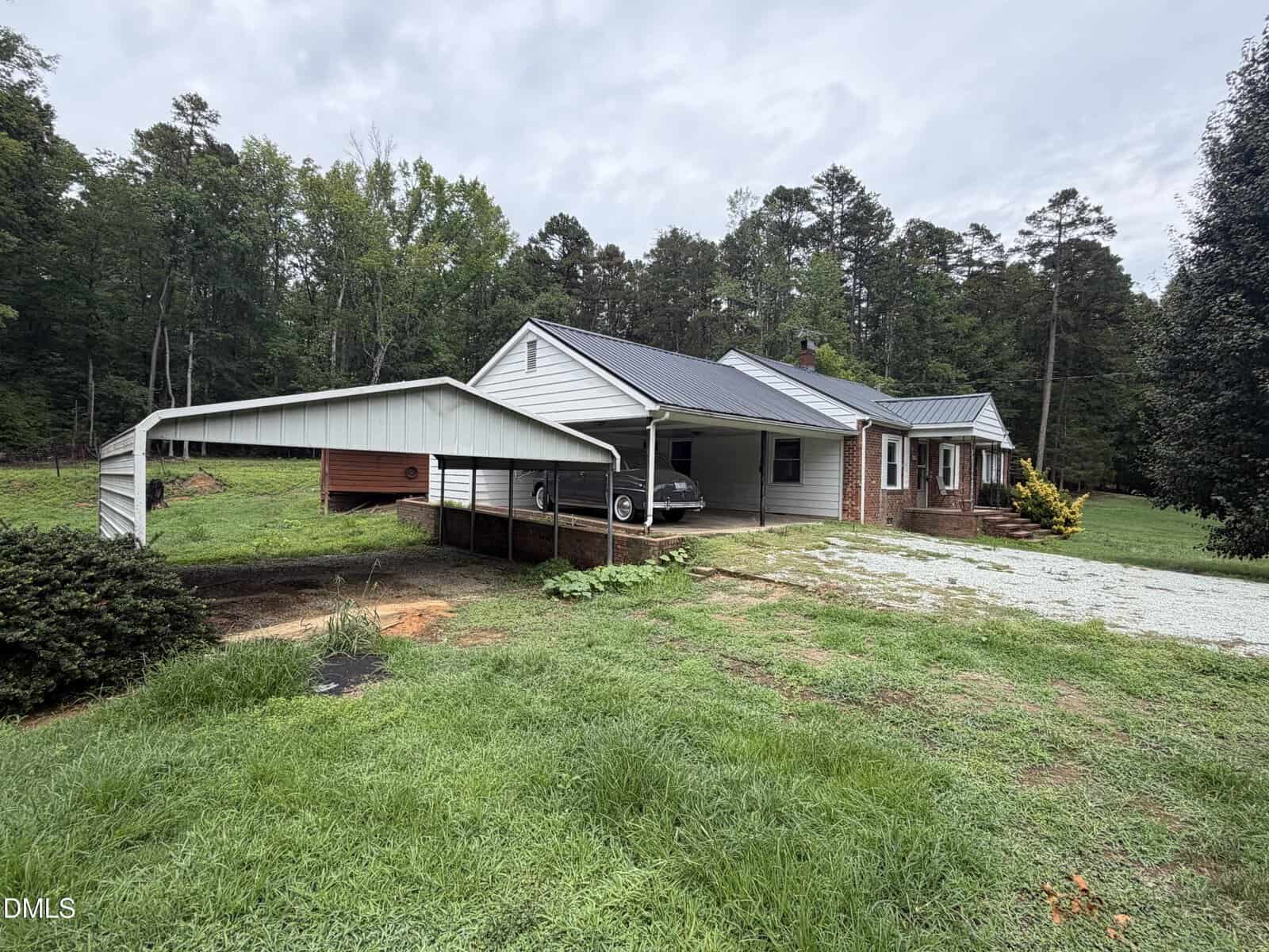 Single-story house with attached carport, a car parked underneath, surrounded by grass and trees, cloudy sky overhead, and gravel driveway leading up to the home.