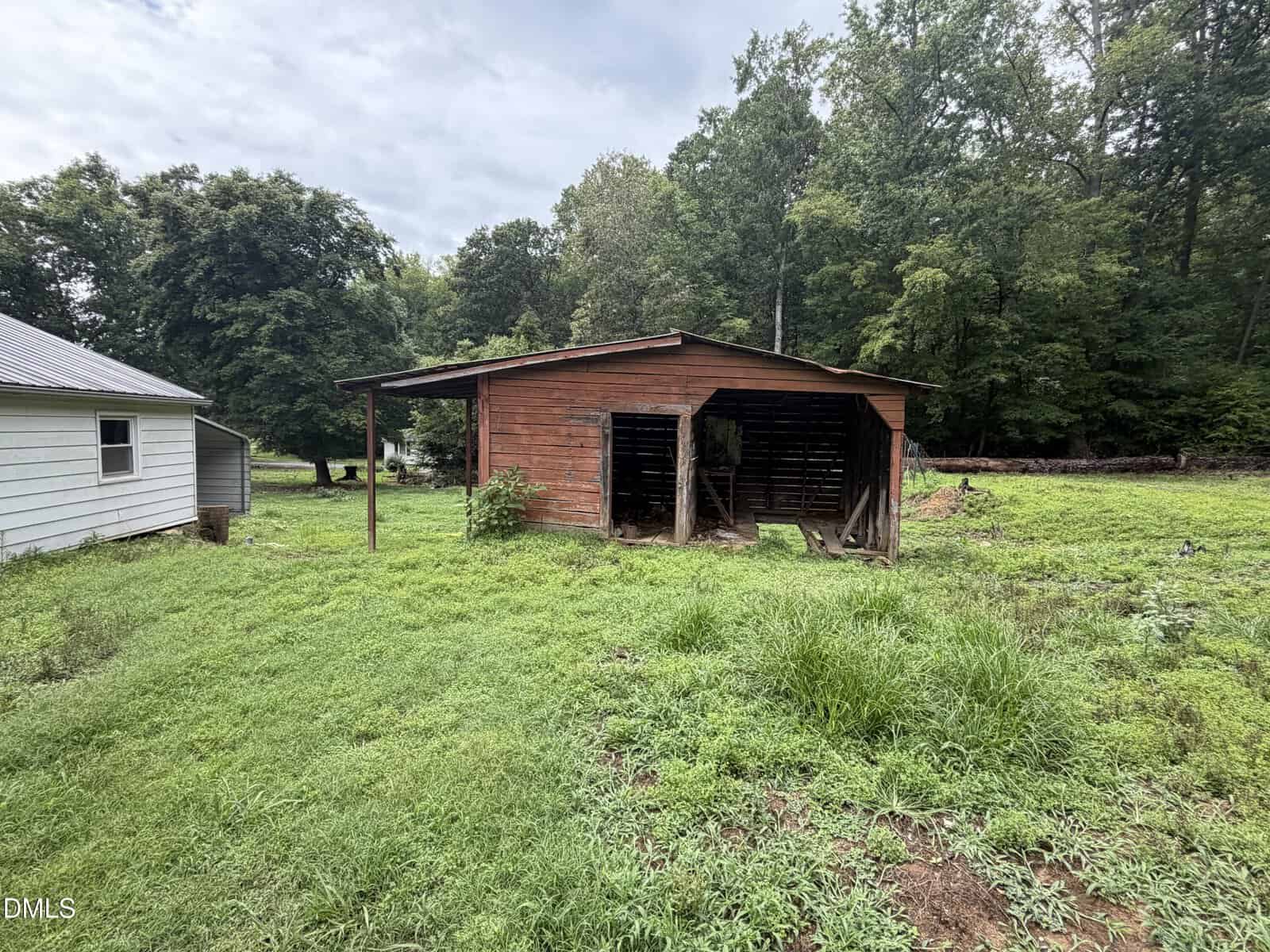 A weathered wooden shed with open sides sits on a grassy lot near a small white building, surrounded by trees under a cloudy sky.
