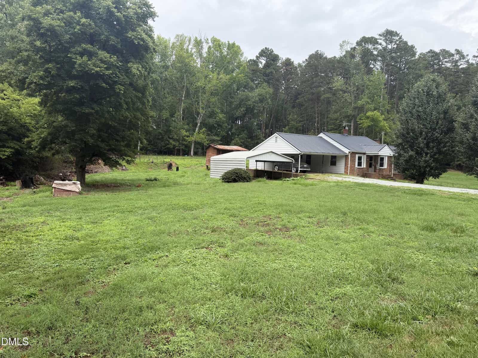 A single-story house with a black roof sits on a large, grassy yard bordered by trees. A driveway leads to a carport beside the house. A shed and a few scattered objects are visible near the edge of the lawn.