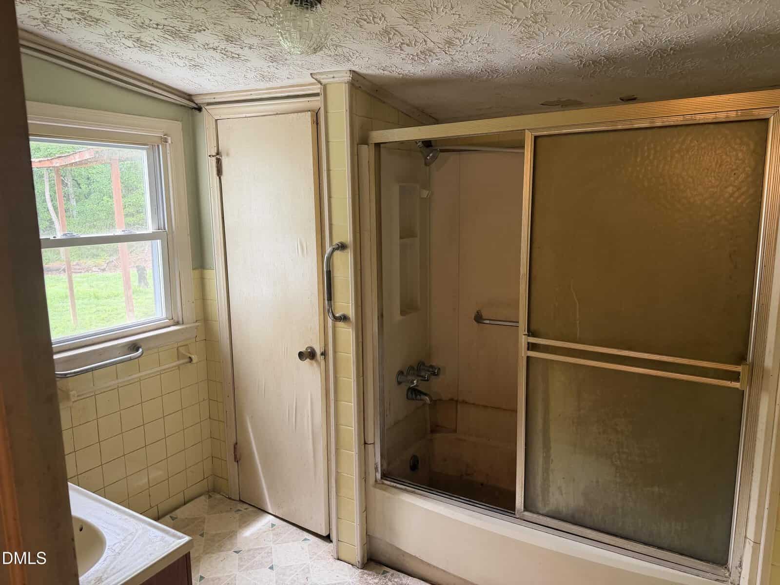 A small, old bathroom with yellowed tiles, a frosted glass shower door, a wooden door, a window letting in daylight, and a partially visible sink. The space appears worn and in need of renovation.