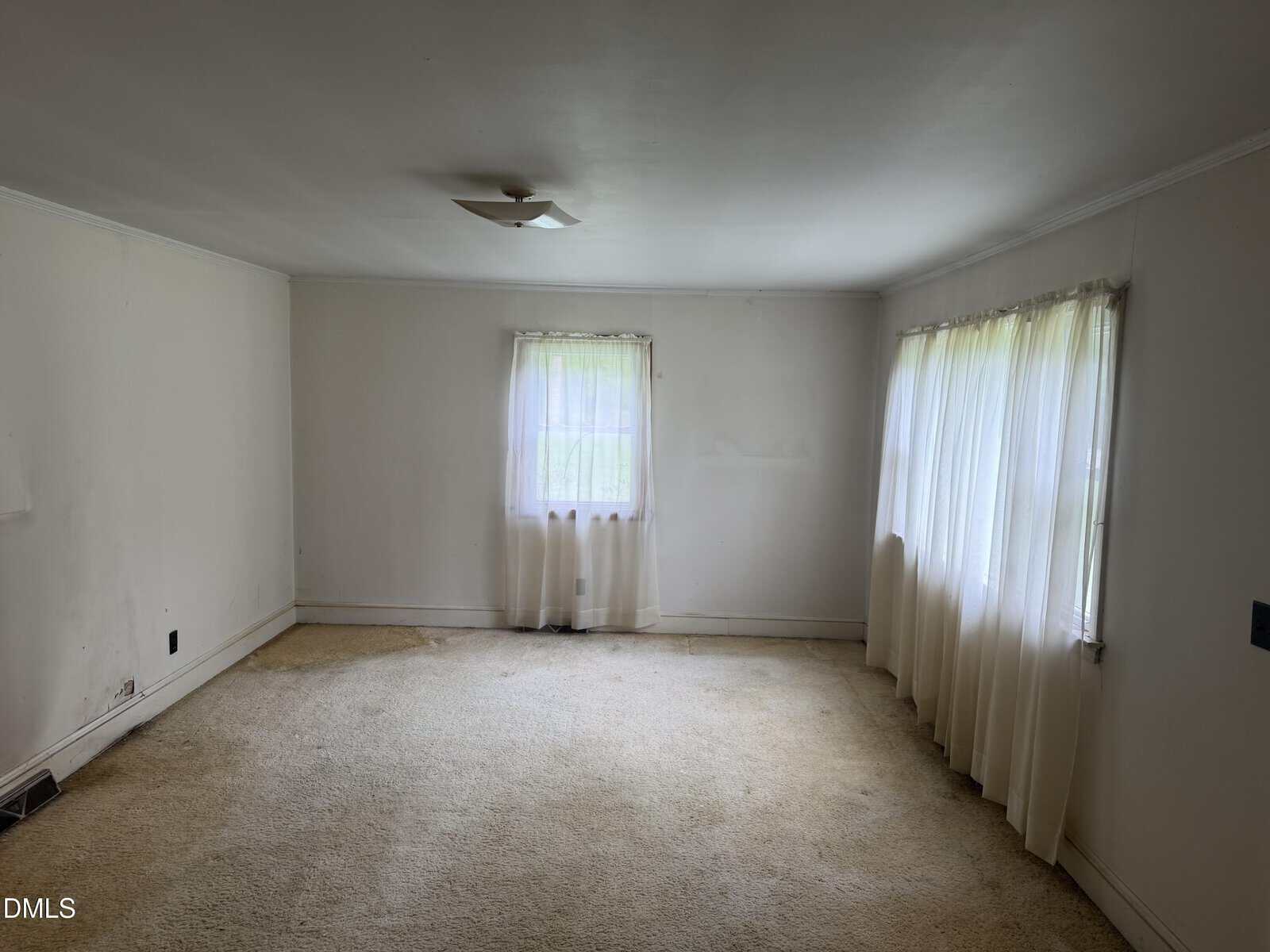 Empty room with off-white walls, beige carpet, two windows with sheer white curtains, and a ceiling light fixture. Natural light fills the space.