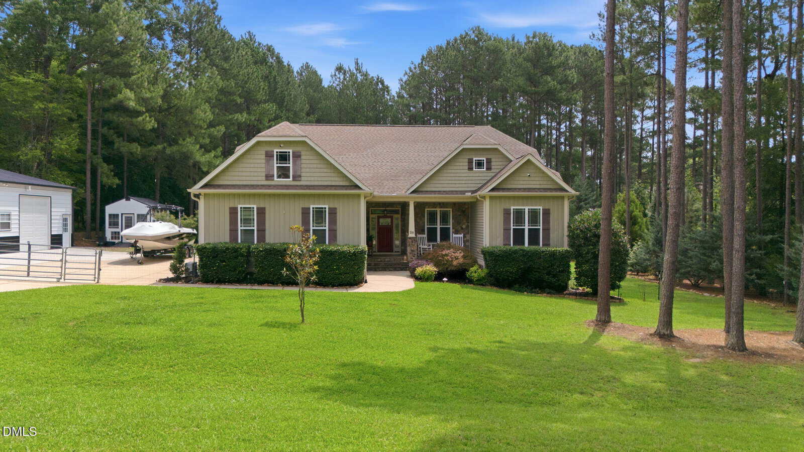 Single-story house with beige siding and green trim, surrounded by a large, neatly mowed lawn, tall trees, and shrubs. A boat is parked on the driveway beside the house, with a forested area in the background.