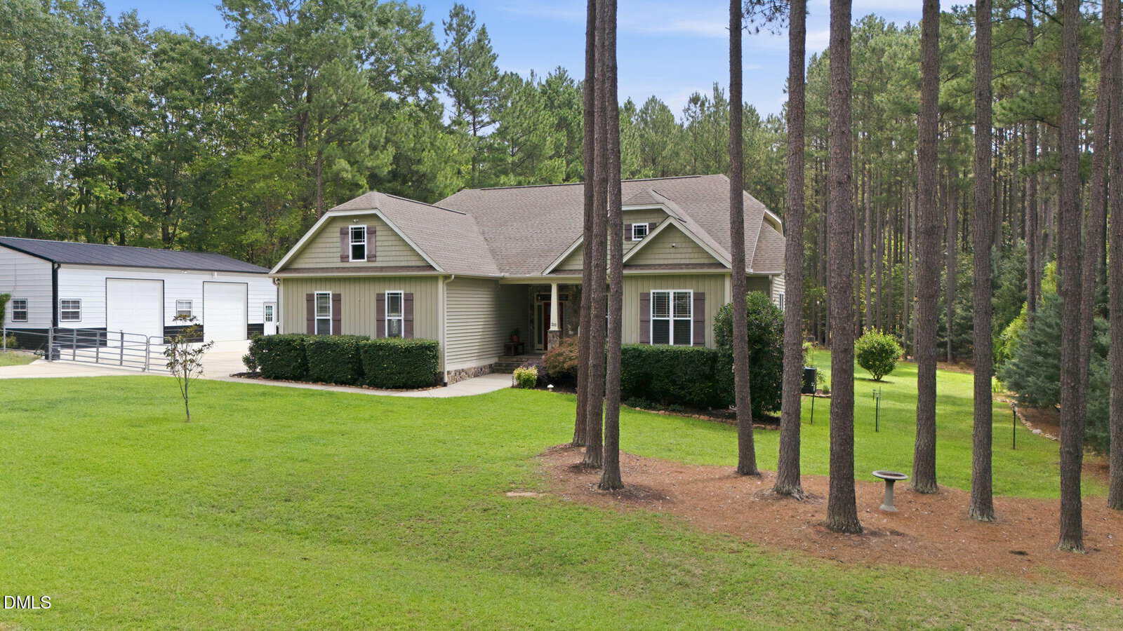 Single-story beige house with white trim, surrounded by neatly trimmed bushes and tall pine trees, sits on a large grassy yard. A white metal garage or workshop is visible to the left, and a birdbath is in the foreground.