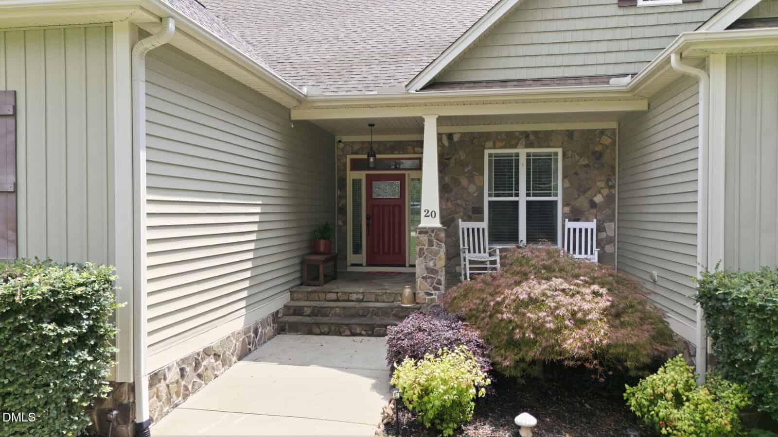 House entrance with red front door, stone accents, two white rocking chairs on the porch, potted plants, shrubs, and a small tree by the walkway. House siding is beige with a gabled roof.