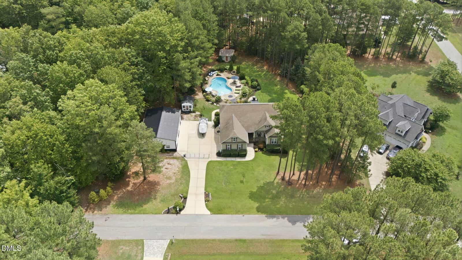 Aerial view of a large house with a circular driveway, swimming pool, gazebo, and surrounding trees. There are neighboring homes and cars on a residential street.