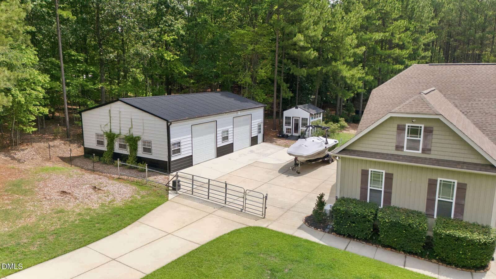 A driveway leads to a large garage with three white doors, a boat on a trailer, a small shed, and a house with green siding, all surrounded by trees and landscaping.