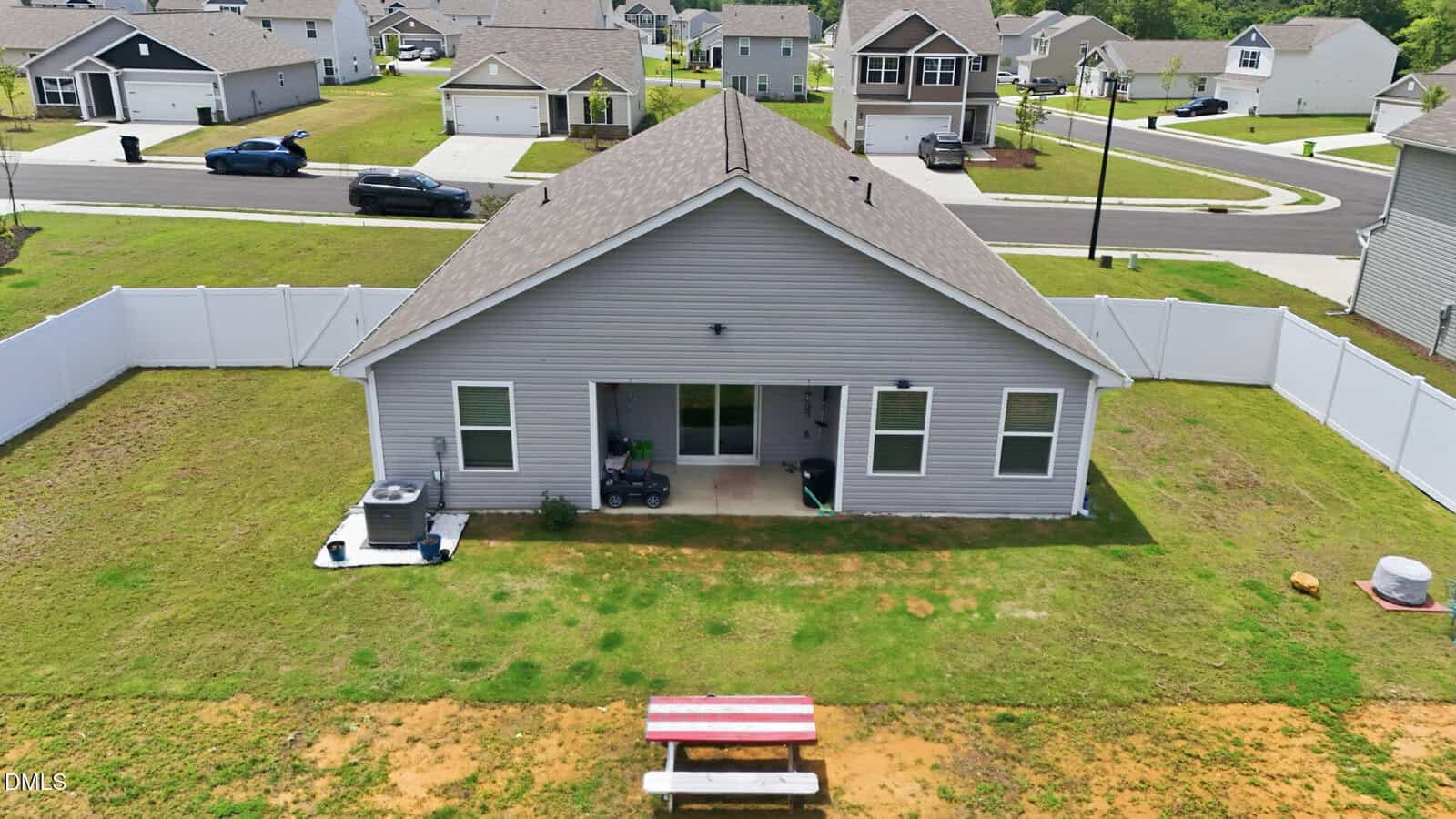 Aerial view of a fenced backyard with a gray house, small patio, central air unit, and a red-and-white picnic table on grass. Suburban homes and streets are visible in the background.