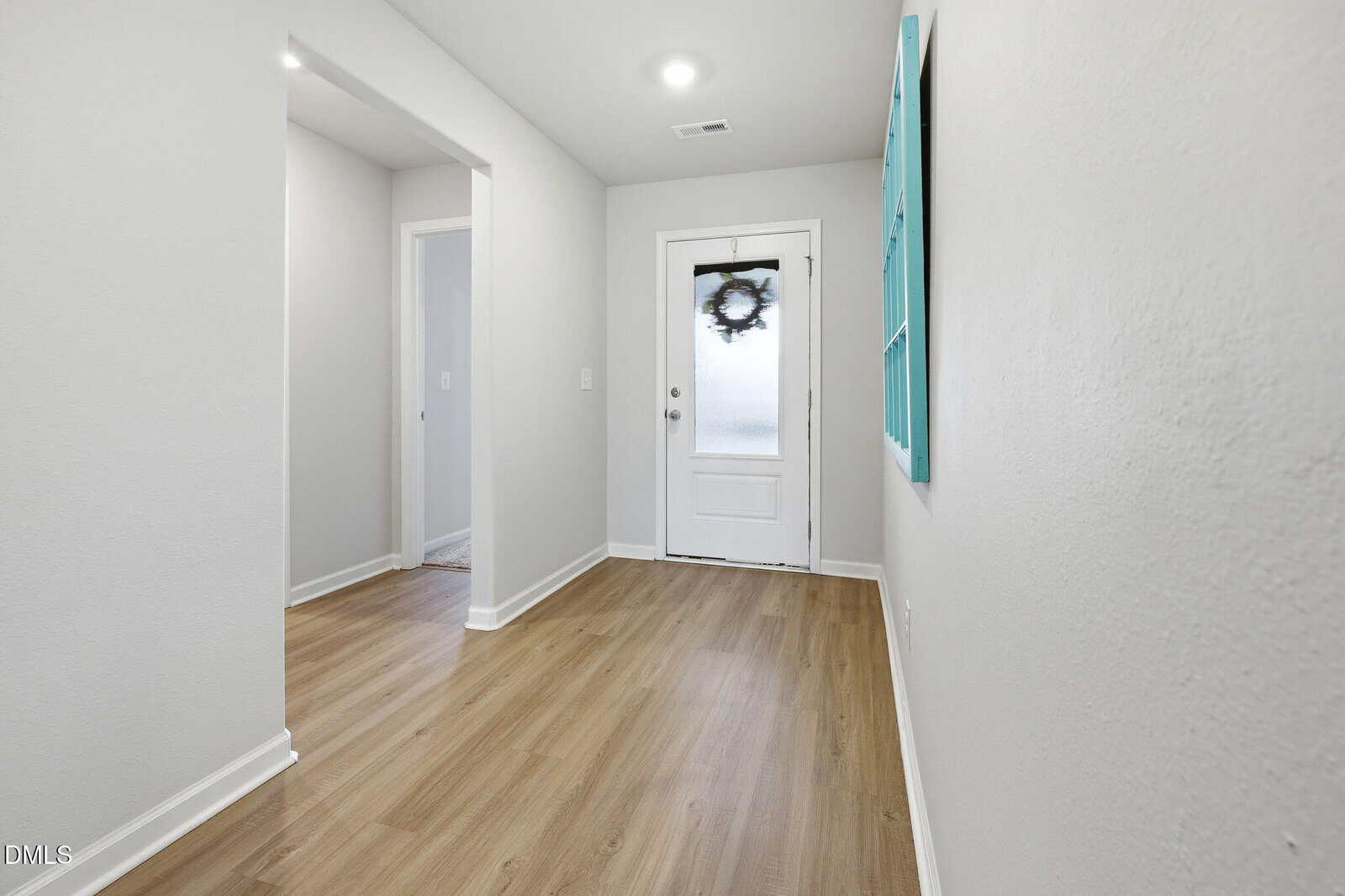 A hallway with light wood flooring, white walls, a door with frosted glass and a wreath, and a large turquoise-framed mirror on the right wall. A doorway on the left leads to another room.