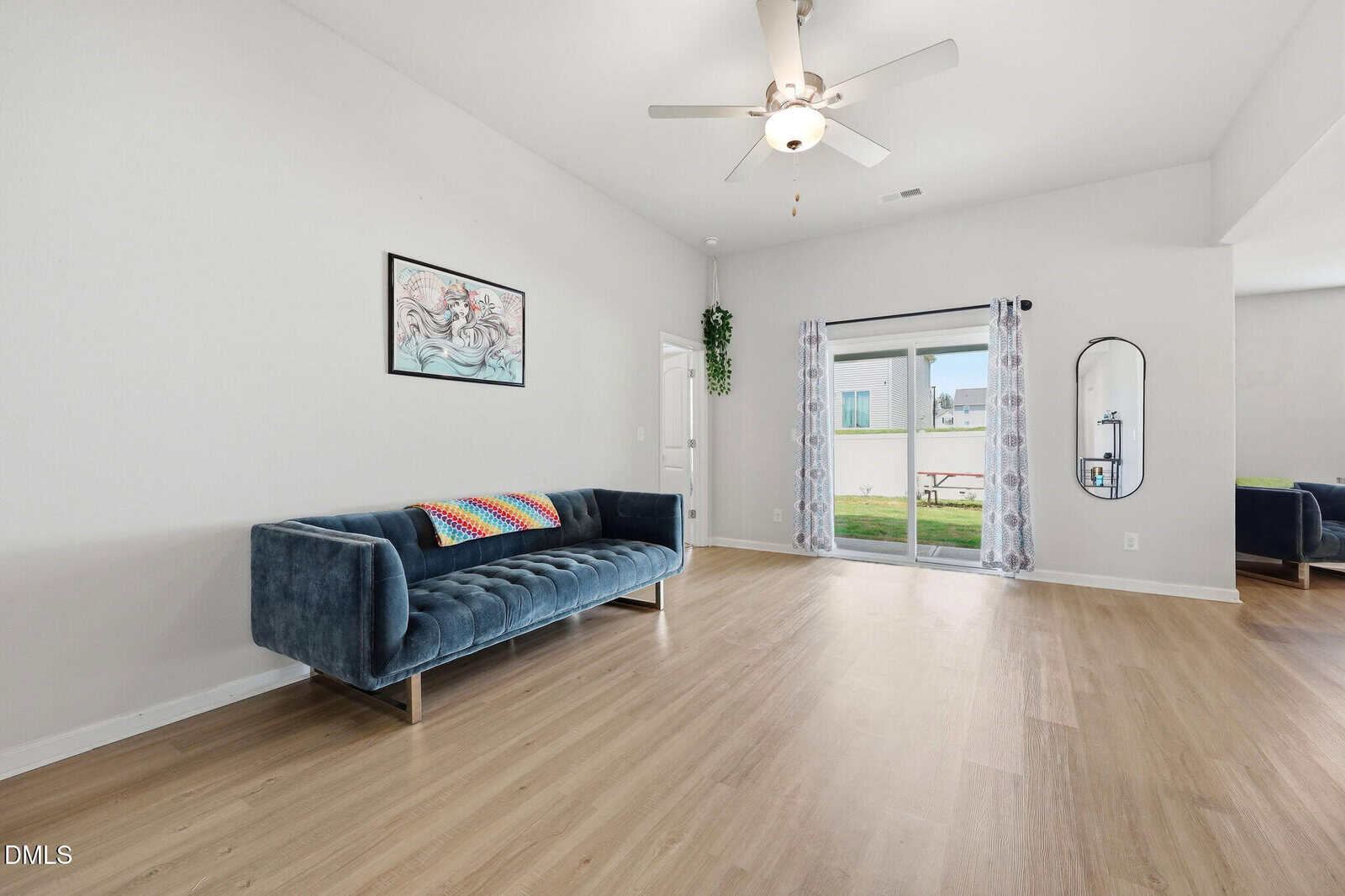 Bright living room with a modern blue sofa, colorful pillow, wall art, ceiling fan, wood floor, glass doors with sheer curtains, and a round mirror; view of green lawn outside.