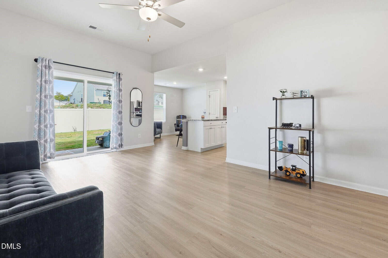Bright living room with light wood floors, a dark couch, sliding glass doors with patterned curtains, a ceiling fan, and a view into a white kitchen. A metal shelf with decor and toys stands against the right wall.