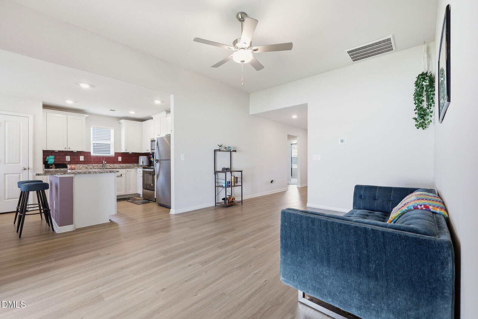 Bright living space with light wood flooring, blue velvet couch, ceiling fan, open kitchen with white cabinets, stainless steel appliances, barstools, and a small shelving unit, all in a modern, minimalist style.