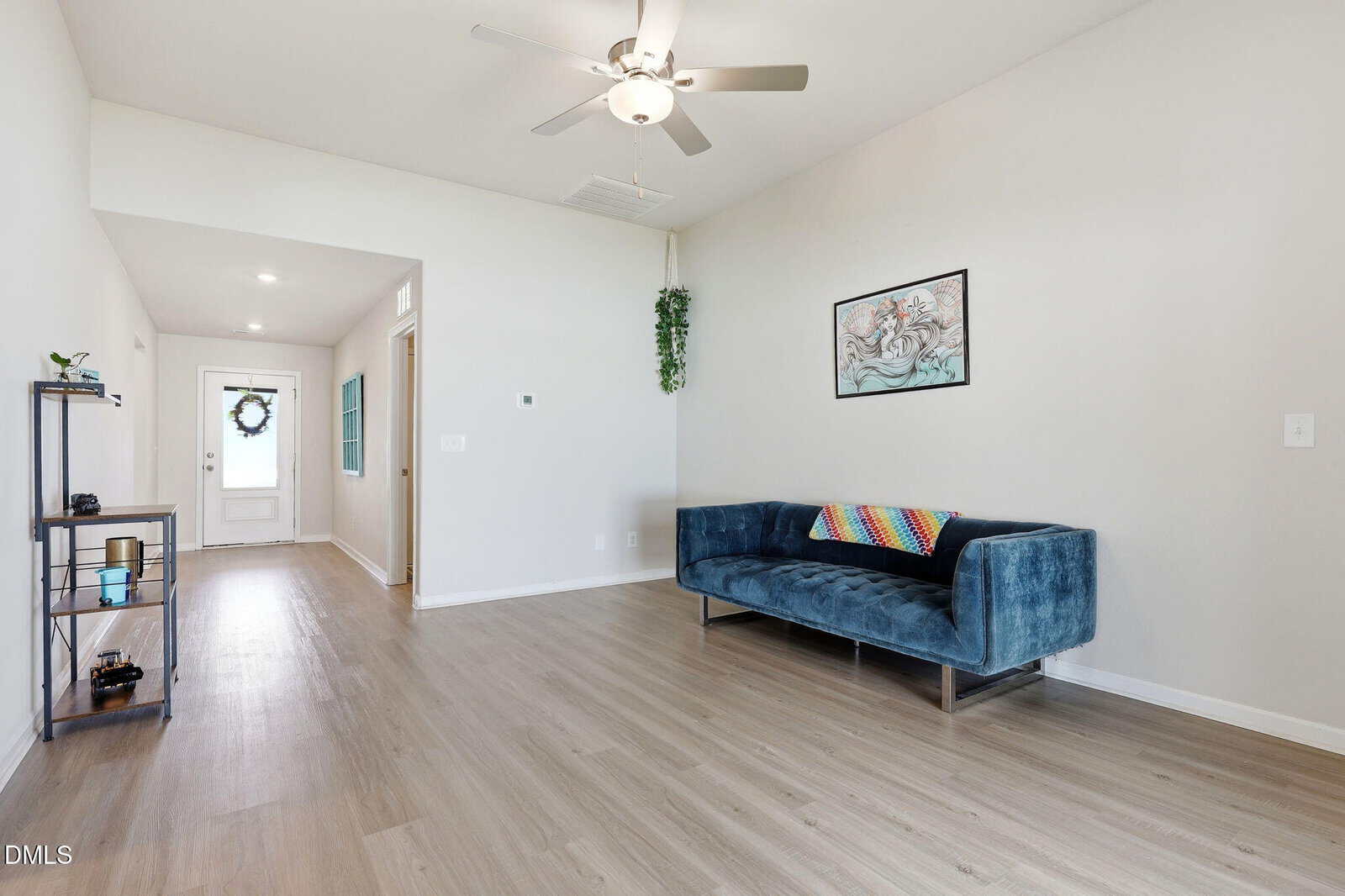 Bright living room with light wood floors, blue velvet sofa with a colorful blanket, wall art, ceiling fan, small shelving unit, and a hallway leading to a door with a wreath.