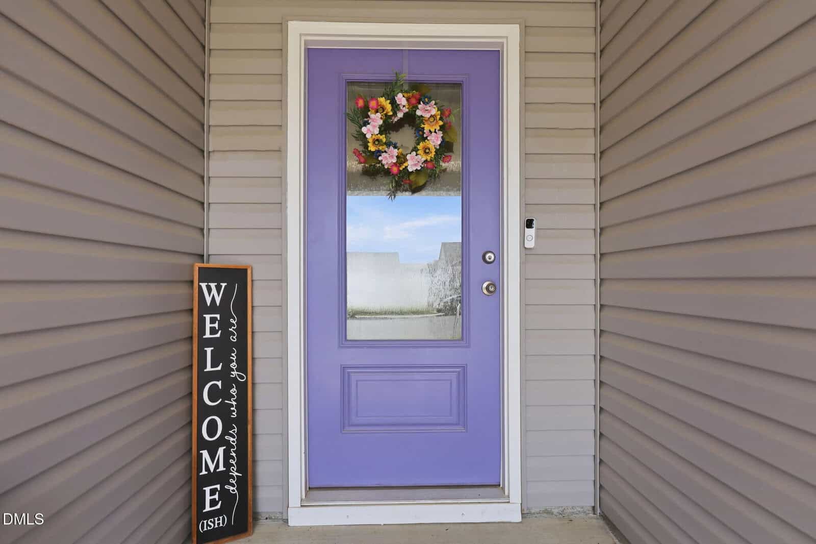 A lavender front door with a floral wreath, a vertical welcome sign leaning beside it, and light gray siding on the house. A reflection of the sky can be seen in the door's window.
