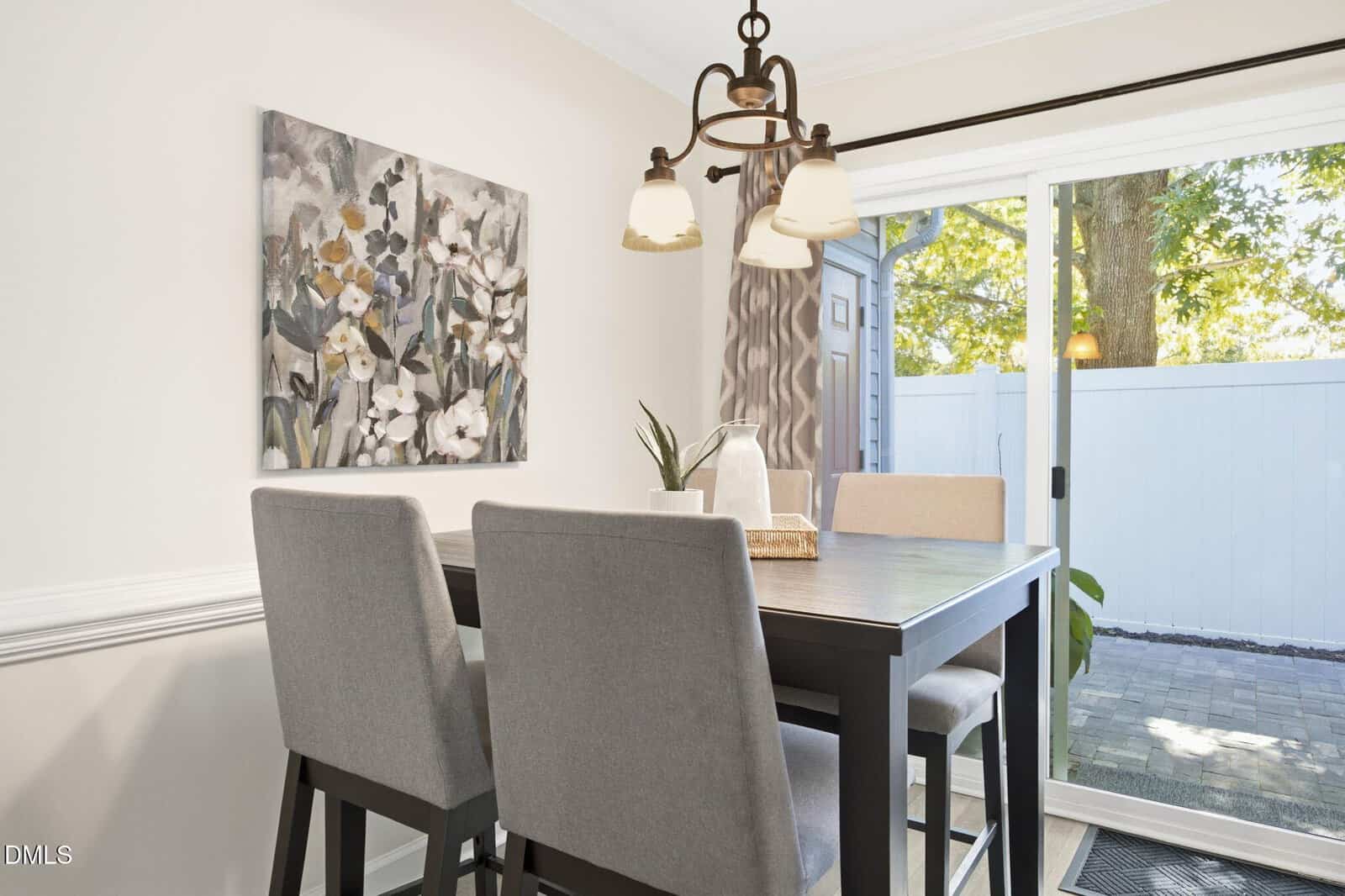 A dining area with a dark wood table, four light gray upholstered chairs, floral wall art, a modern light fixture, and a sliding glass door leading to a patio with trees and a white fence outside.