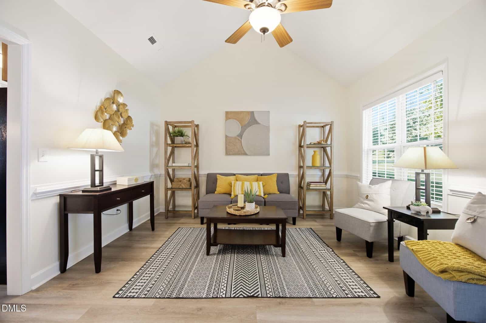 Bright living room with a gray sofa, yellow accents, wooden shelves, armchairs, a patterned rug, ceiling fan, and large windows letting in natural light. Decor includes abstract wall art and plants.