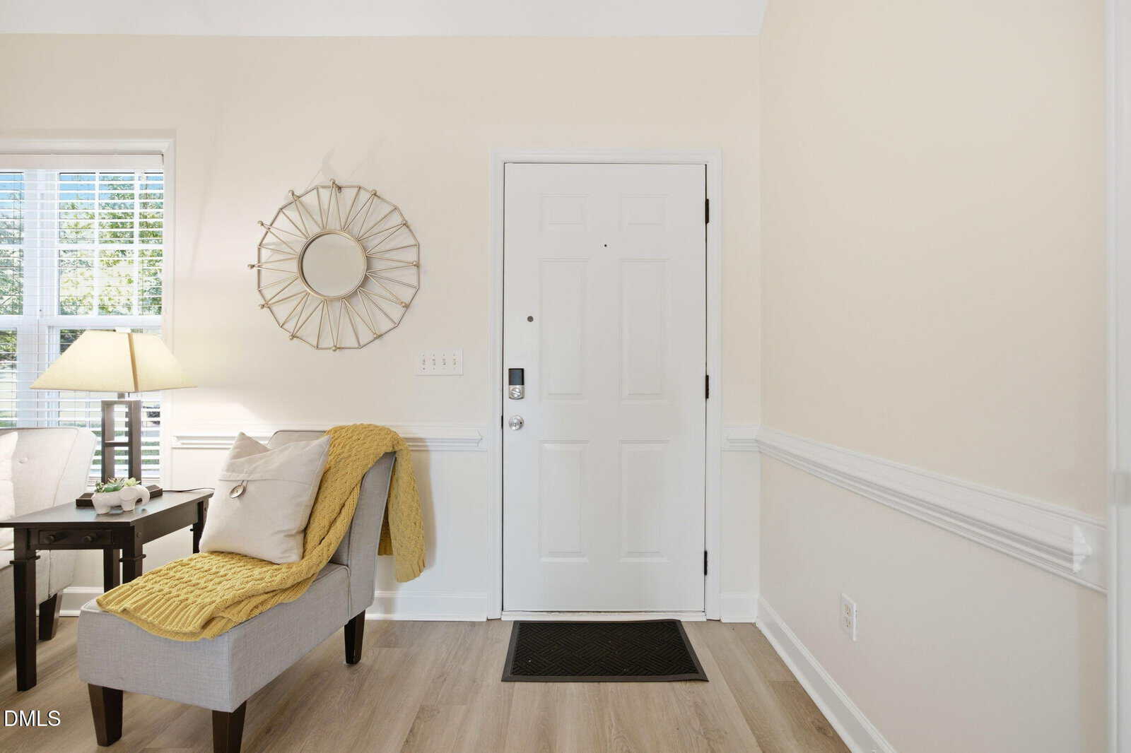A bright entryway with a white front door, beige walls, a light gray chair with a yellow throw, a small table with a lamp, and a decorative mirror above light wood floors.