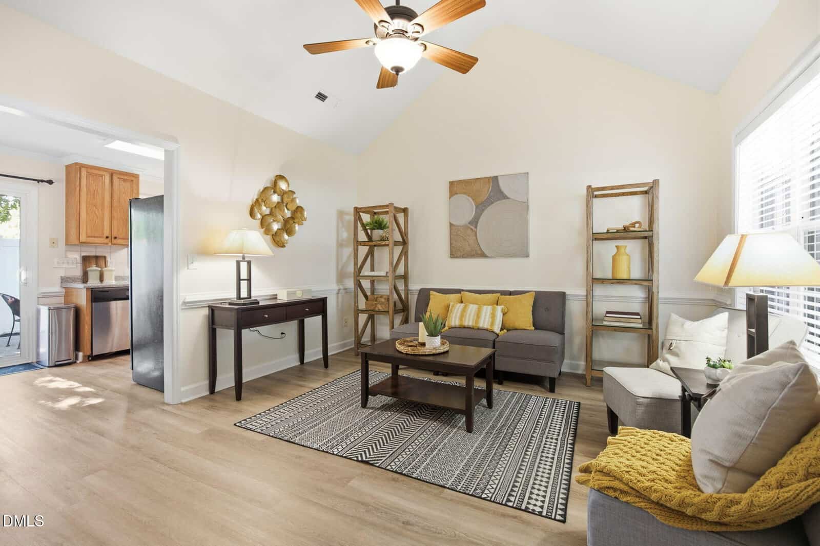Bright living room with a ceiling fan, gray sofa, armchair, bookshelves, coffee table, and area rug. Art and plants decorate the space. Sunlight streams in from large windows. Kitchen is visible through an open doorway.