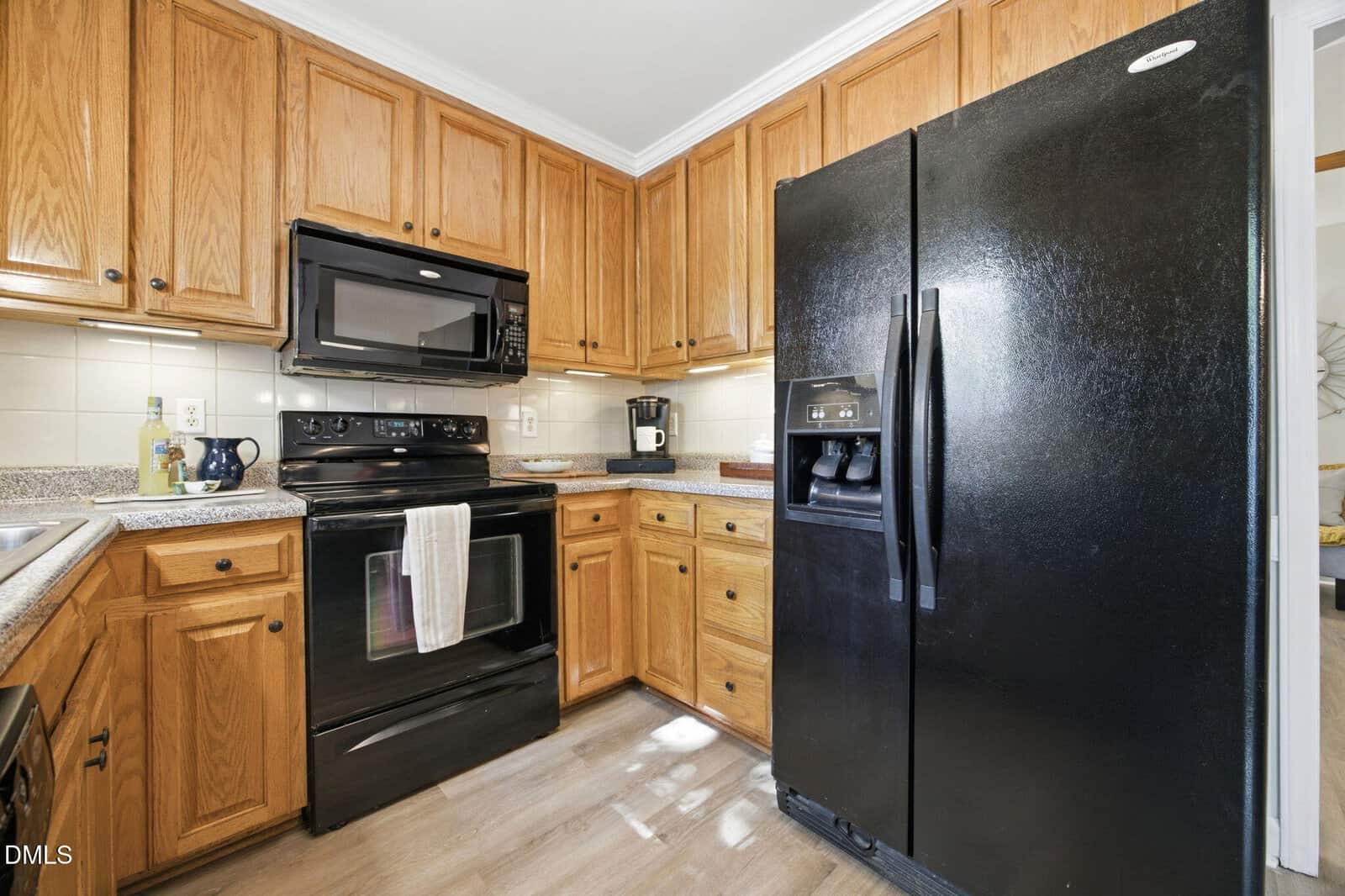 A kitchen with wooden cabinets, black appliances including a refrigerator, stove, microwave, and a coffee maker on the counter. A white dish towel hangs on the oven door. Light wood flooring is visible.