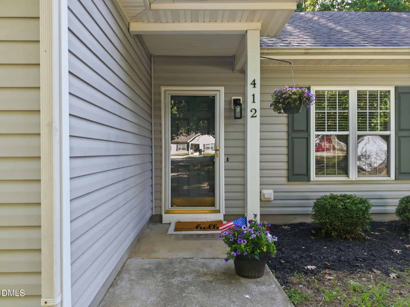 Gray house front porch with the number 412, a glass storm door, a doormat that says "hello," potted flowers with small American flags, a hanging flower basket, and two windows with green shutters.