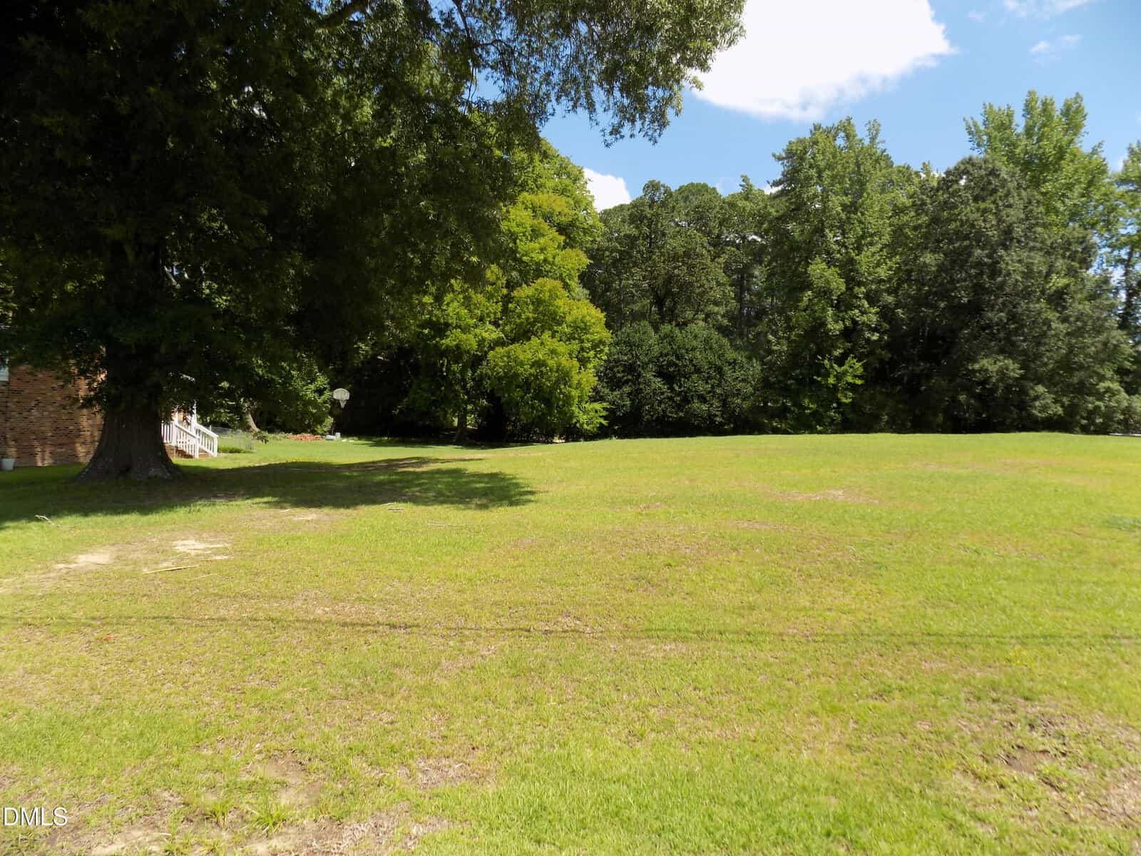 Grassy open field with scattered trees bordering the area, under a partly cloudy sky. A house is partially visible on the left edge, mostly obscured by a large tree.