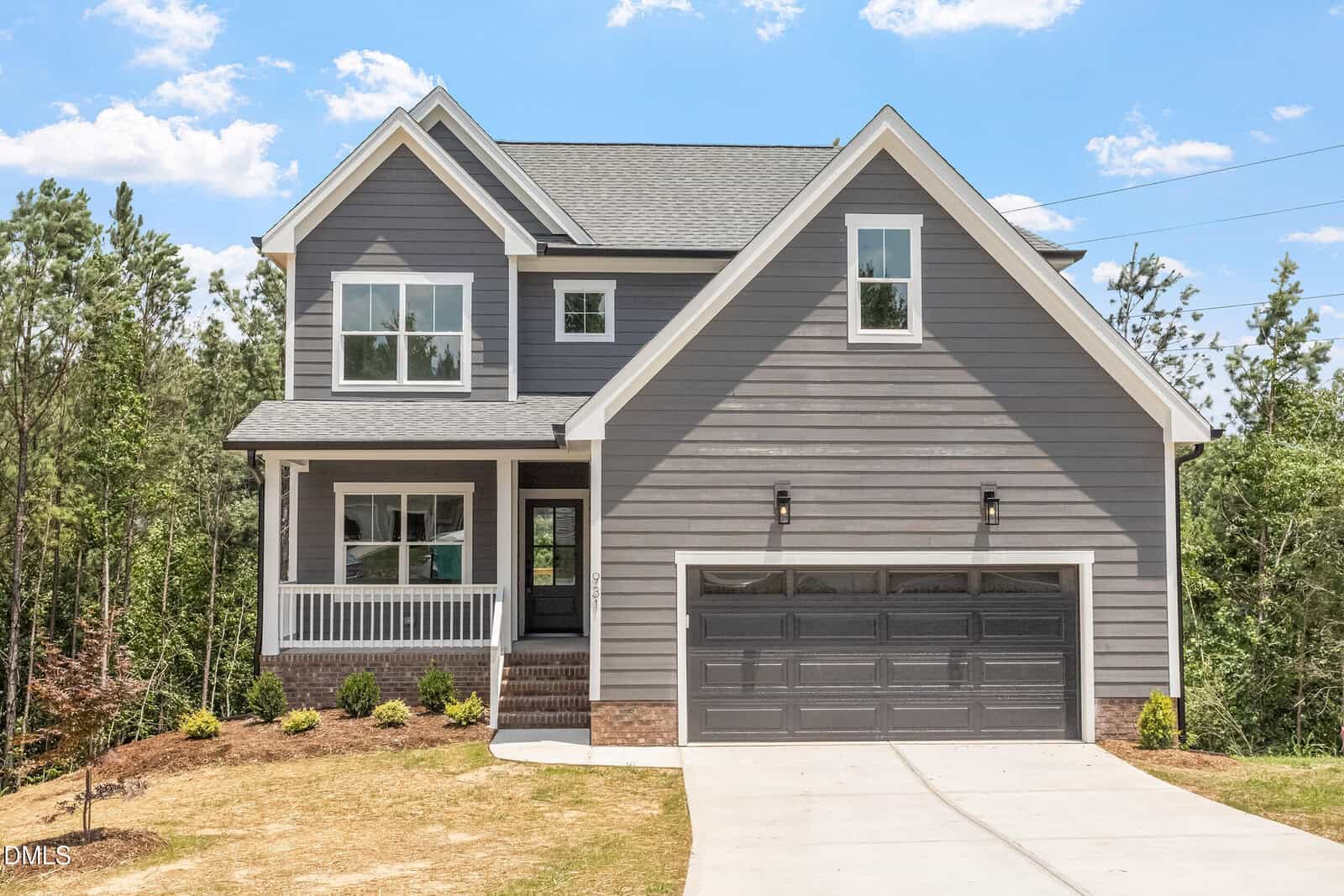 Charming two-story gray house with white trim located at 931 Woodland Road in Creedmoor. Featuring a welcoming front porch, an attached two-car garage, and a concrete driveway, this home offers both style and convenience. The yard includes minimal landscaping with small shrubs and brown grass, while mature trees provide a scenic backdrop.