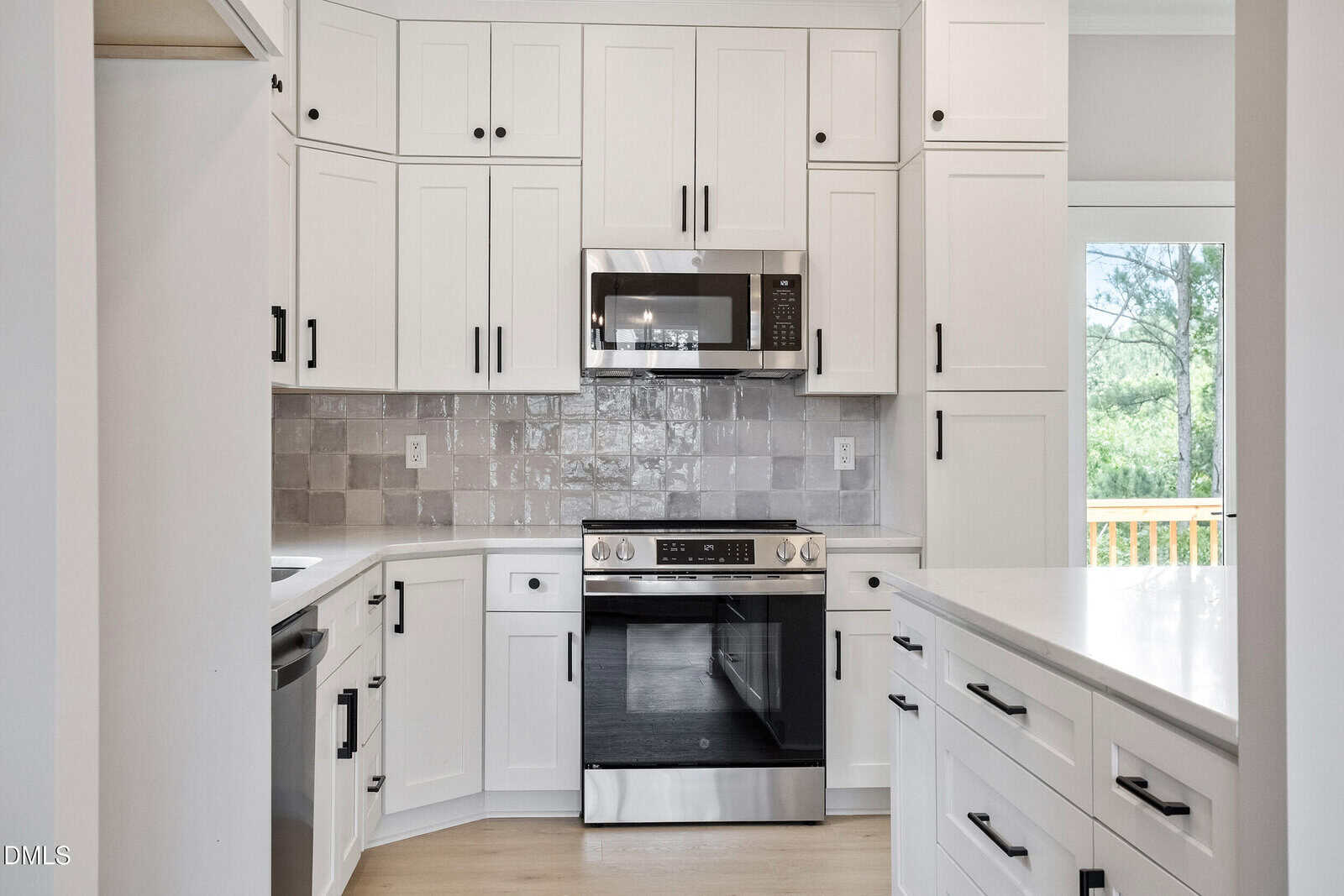 Modern kitchen with white cabinets, stainless steel oven and microwave, light gray tile backsplash, and large windows letting in natural light. Black hardware accents the cabinetry throughout the space.