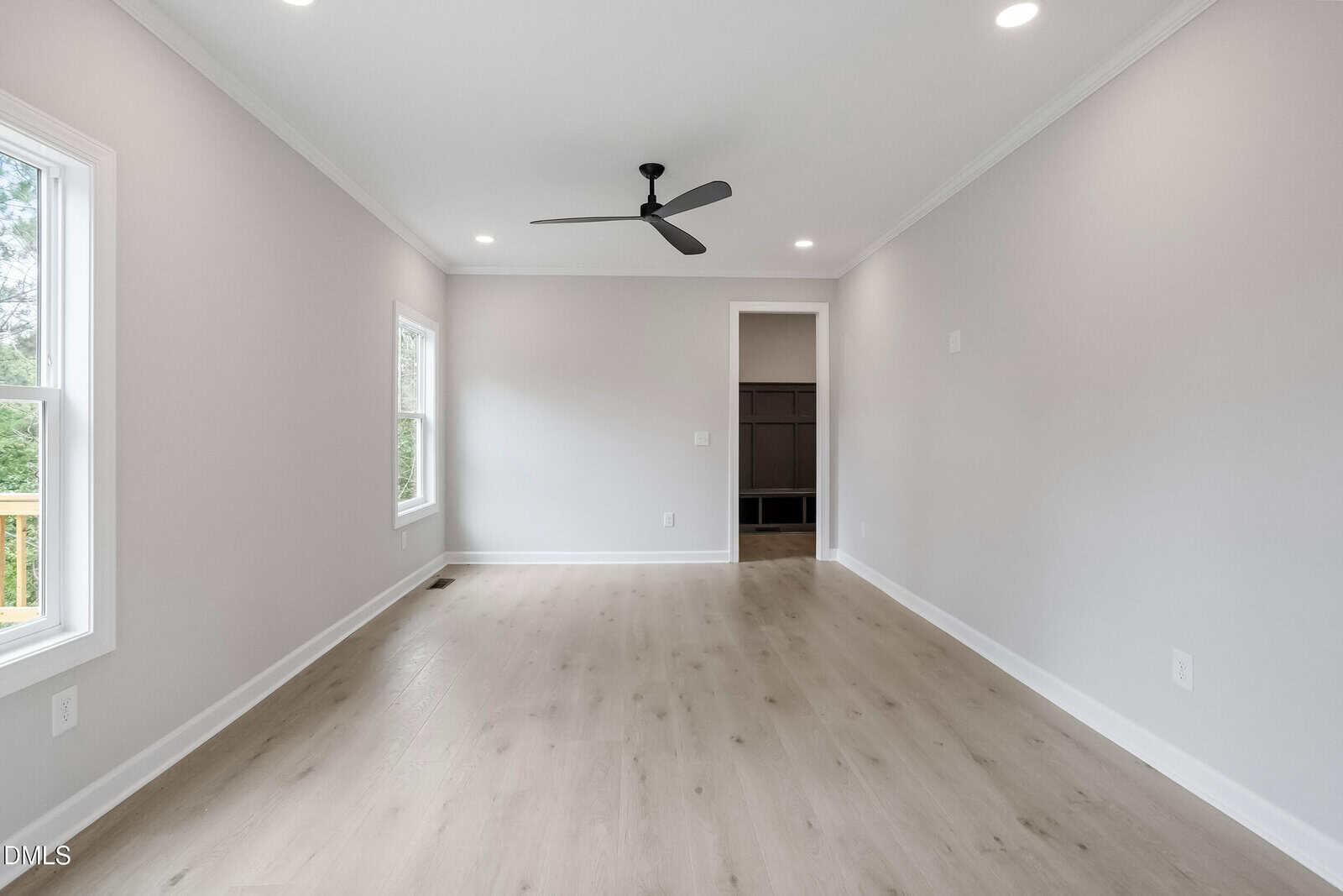 A bright, empty room with light wood floors, white walls, recessed ceiling lights, a ceiling fan, and two windows. An open doorway leads to another space.