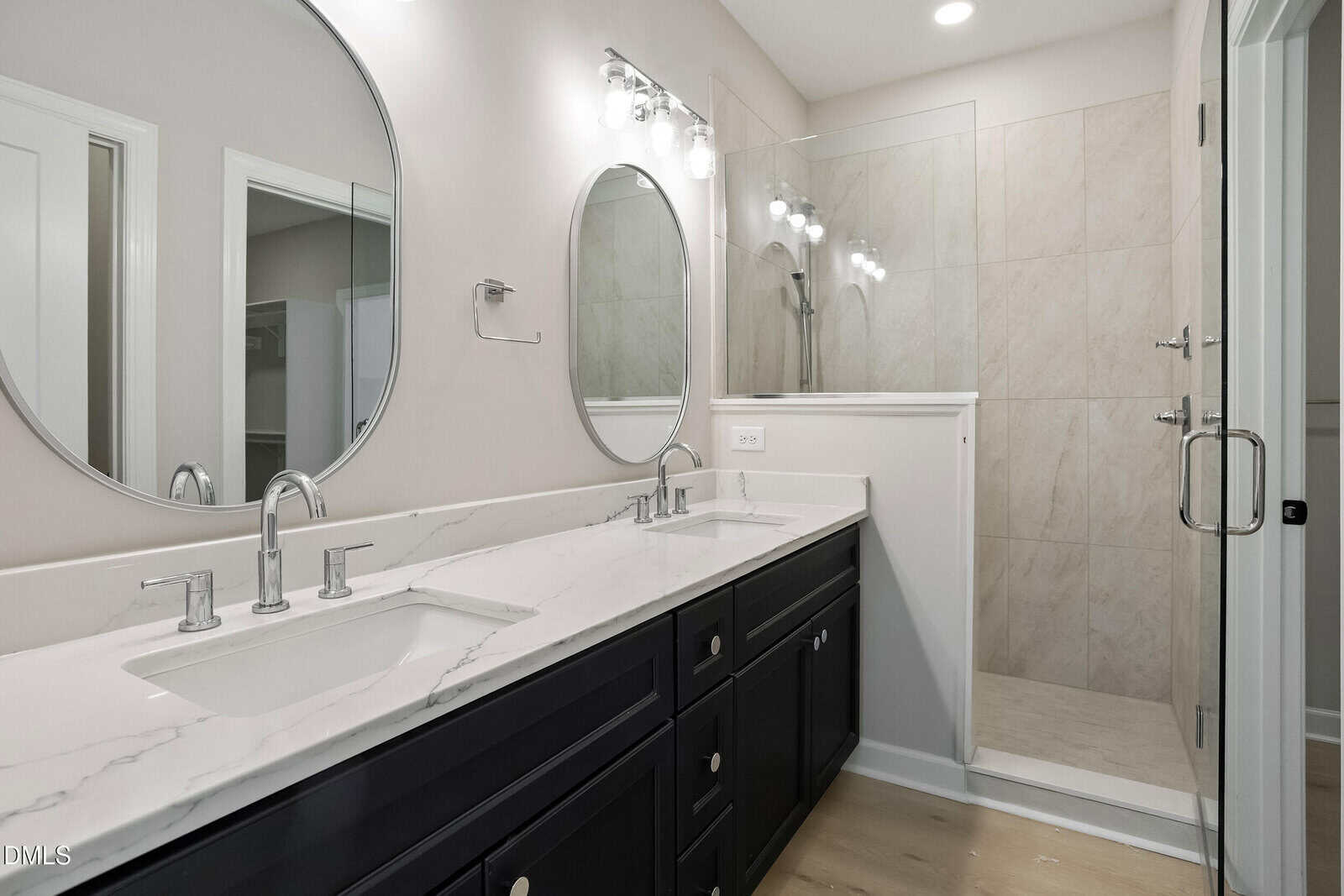 Modern bathroom with double sinks, marble countertop, oval mirrors, dark cabinets, and a glass-door shower with beige tile walls. Bright lighting and neutral-colored walls create a clean, elegant look.