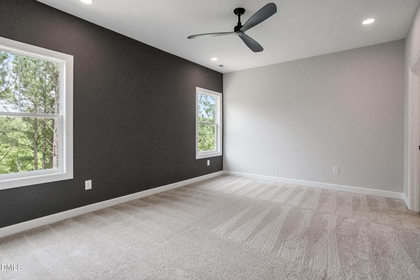 A modern, empty bedroom with light carpet, a dark accent wall, two large windows, a ceiling fan, and recessed lighting. Natural light fills the room, highlighting the clean and simple design.