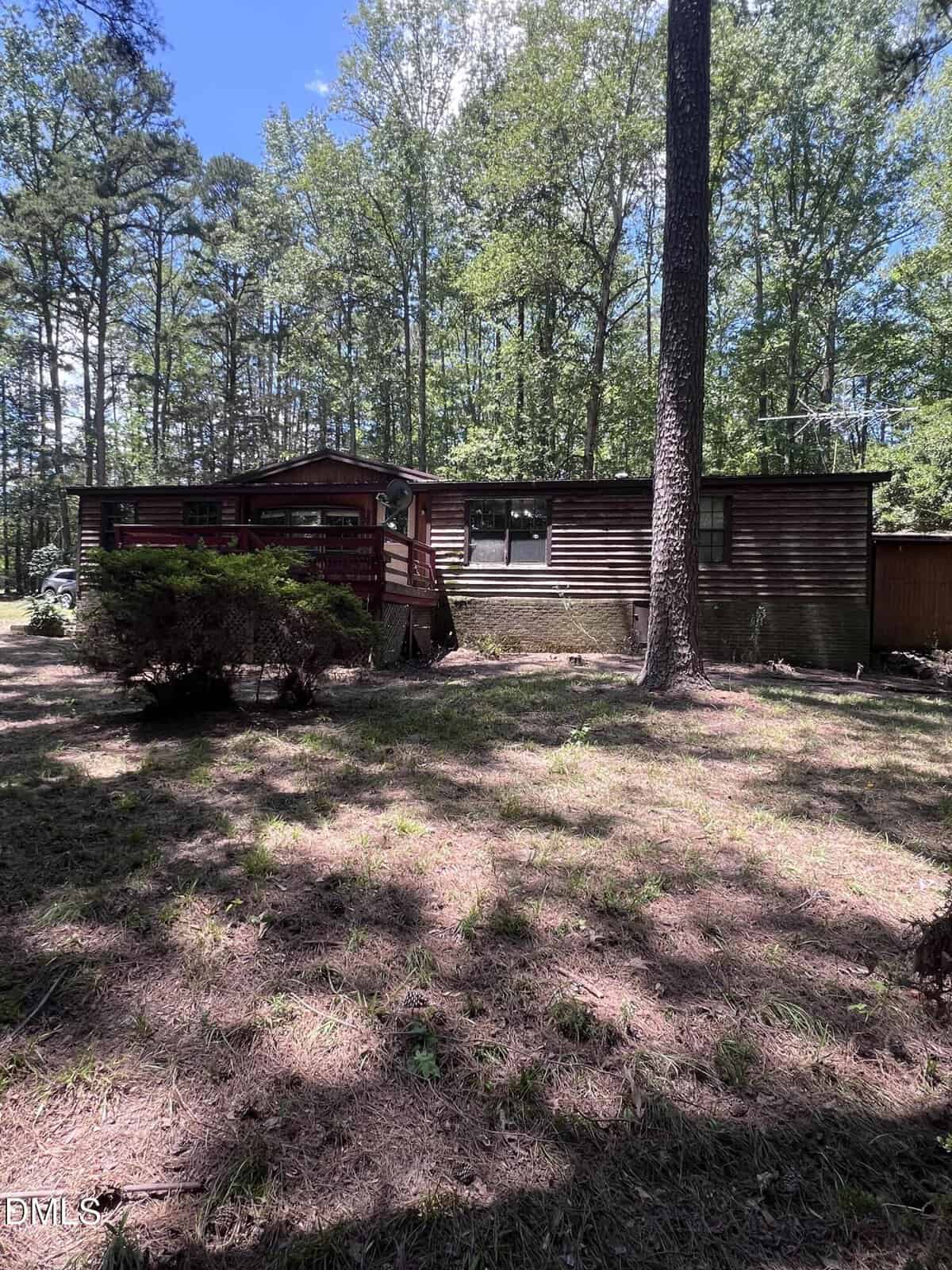 A rustic, single-story wooden cabin with a red deck sits in a sunlit, wooded area on 197 E Holly Lane, Henderson, surrounded by tall trees and patches of grass.