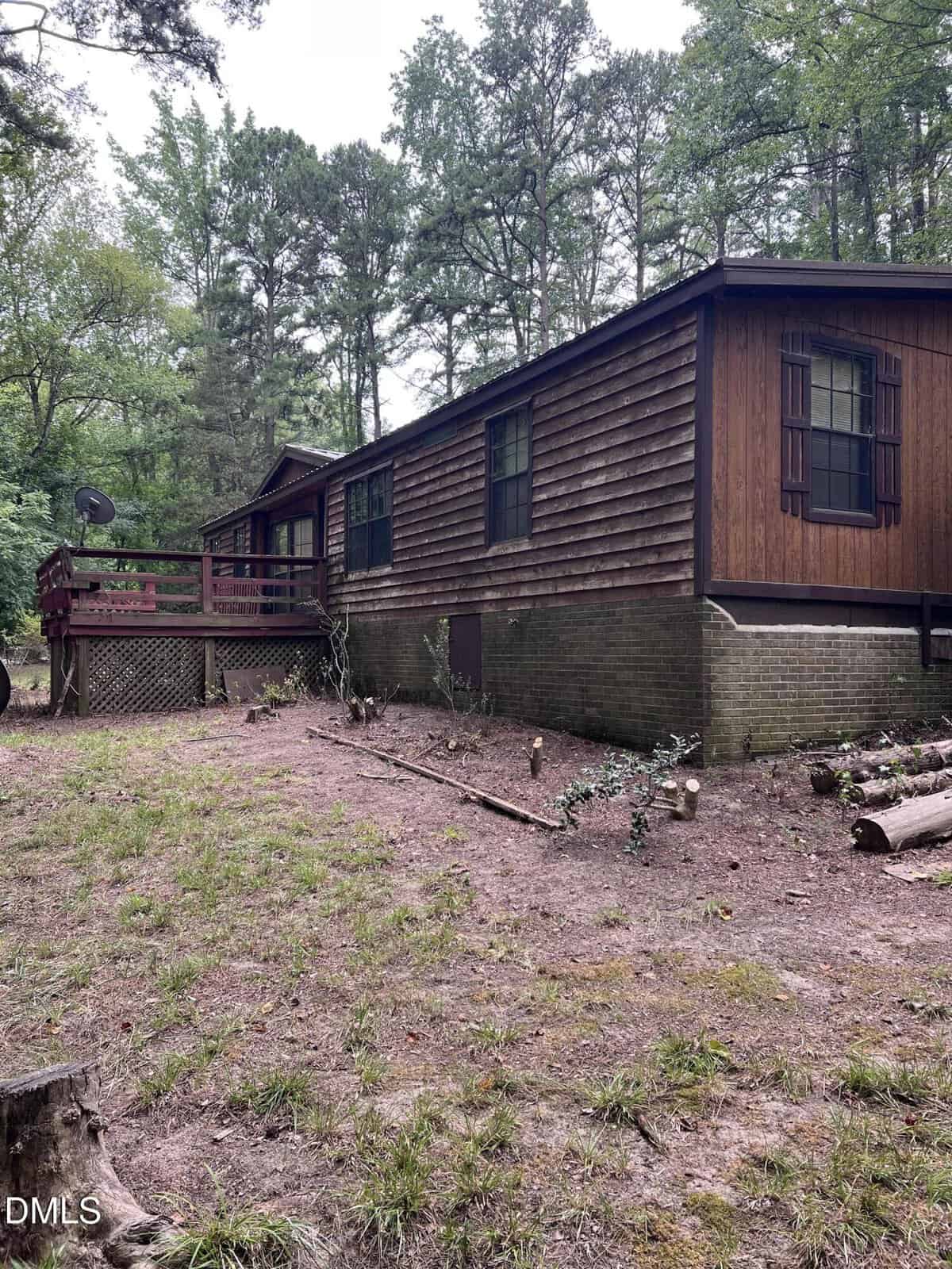 A rustic, single-story wooden cabin with a raised deck sits in a wooded area. The yard features patchy grass and scattered logs, while tall trees surround the house.