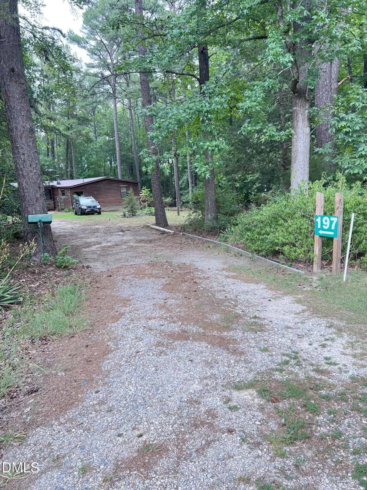 A gravel driveway leads to a wooden house surrounded by tall trees. A green mailbox is on the left, and a sign with the number 197 is posted on the right side of the driveway.