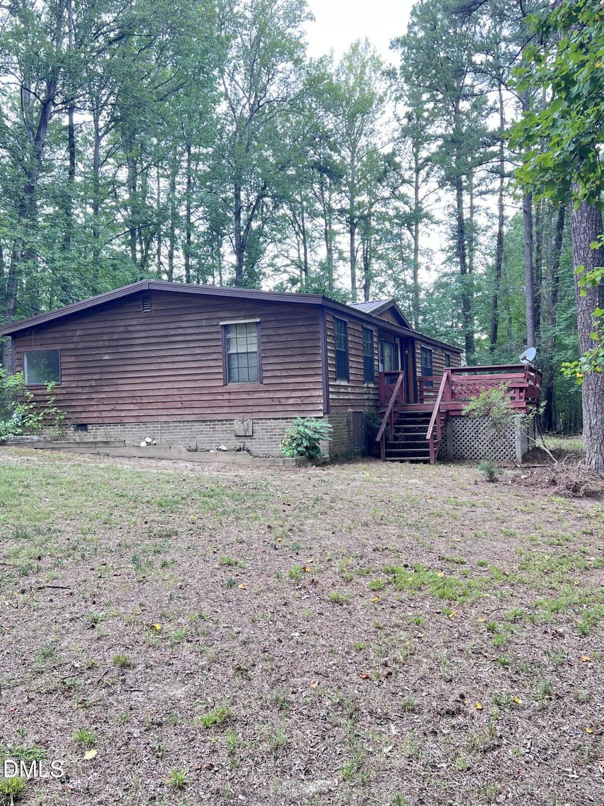 A wooden cabin with a red deck sits among tall trees in a forested area. The ground is covered with sparse grass and pine needles. Steps lead up to the deck on the right side of the house.