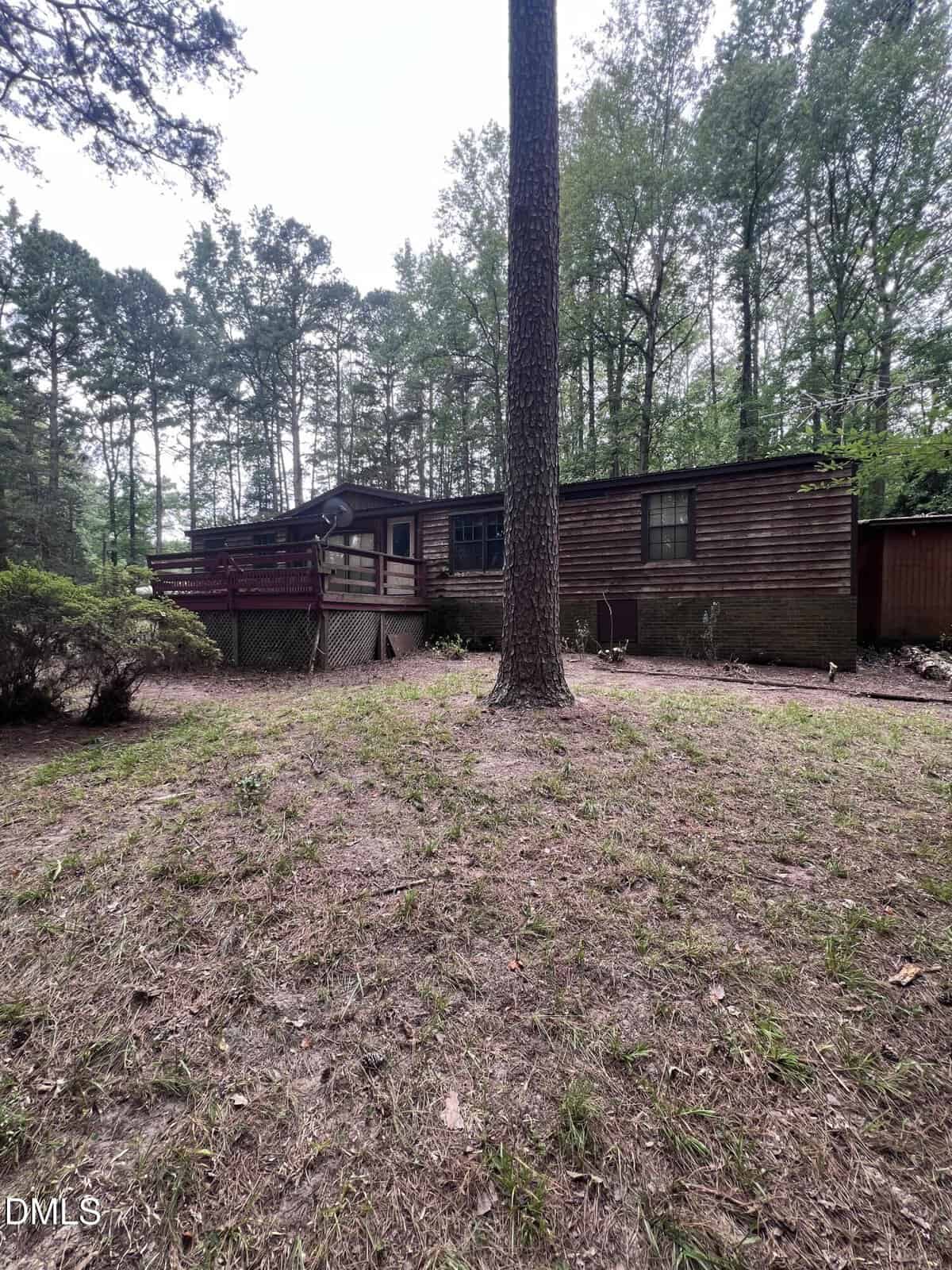 A wooden, single-story house with a deck, surrounded by tall trees and a dirt yard. The home has dark trim and a slanted roof, with satellite dish visible on the deck railing.