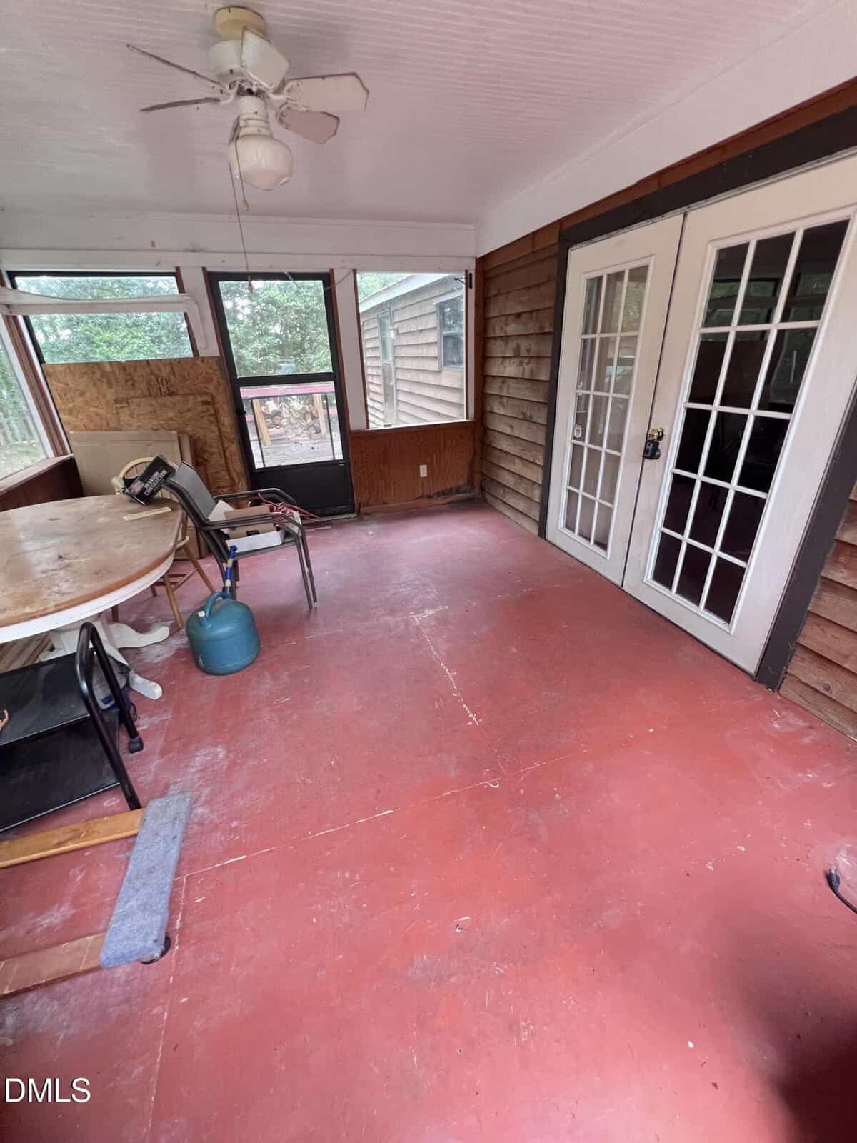 Enclosed porch with red floor, white ceiling fan, round table with chairs, propane tank, and double French doors. Wood paneling lines the walls, and windows provide a view outside.