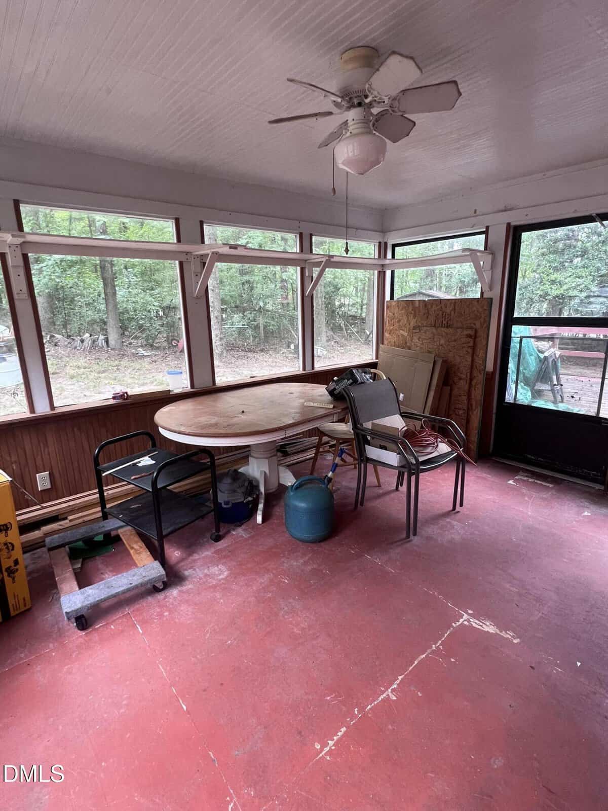 A screened porch with red floor, a ceiling fan, round wooden table, scattered chairs, and miscellaneous items. Large windows show trees and a wooded area outside.