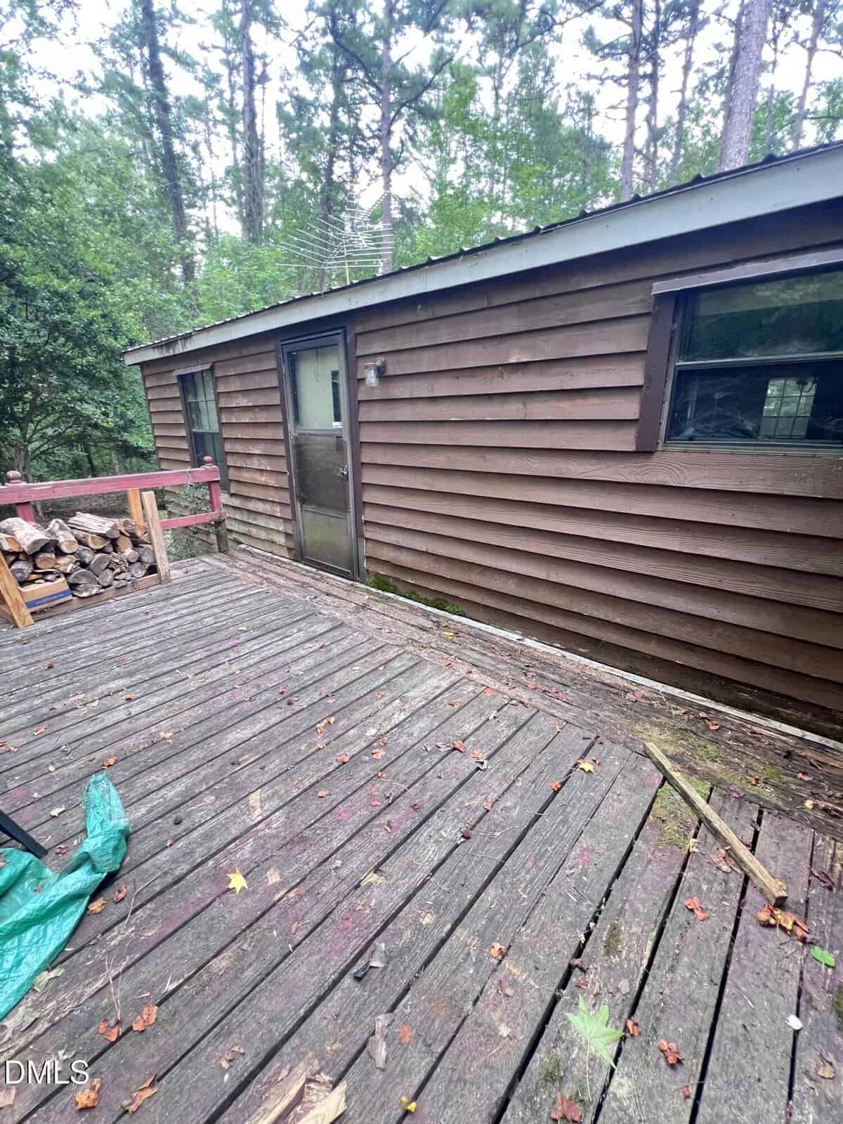 A weathered wooden deck leads to a brown cabin with a metal roof, glass door, and window, surrounded by tall trees and stacked firewood.