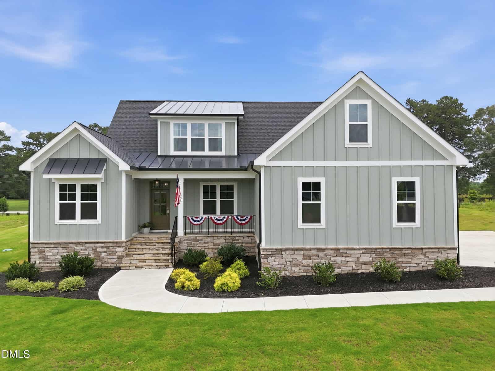 A modern gray house with white trim, stone accents, and a covered front porch decorated with patriotic bunting sits proudly at 35 Wheatfield Lane in Youngsville. The yard is beautifully landscaped with shrubs and a curved sidewalk, all beneath a clear blue sky.