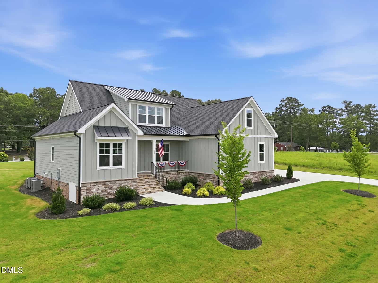 A modern light gray house with dark roof, front porch decorated with patriotic bunting, stone steps, landscaped yard, and a curved driveway on a green lawn under a blue sky.