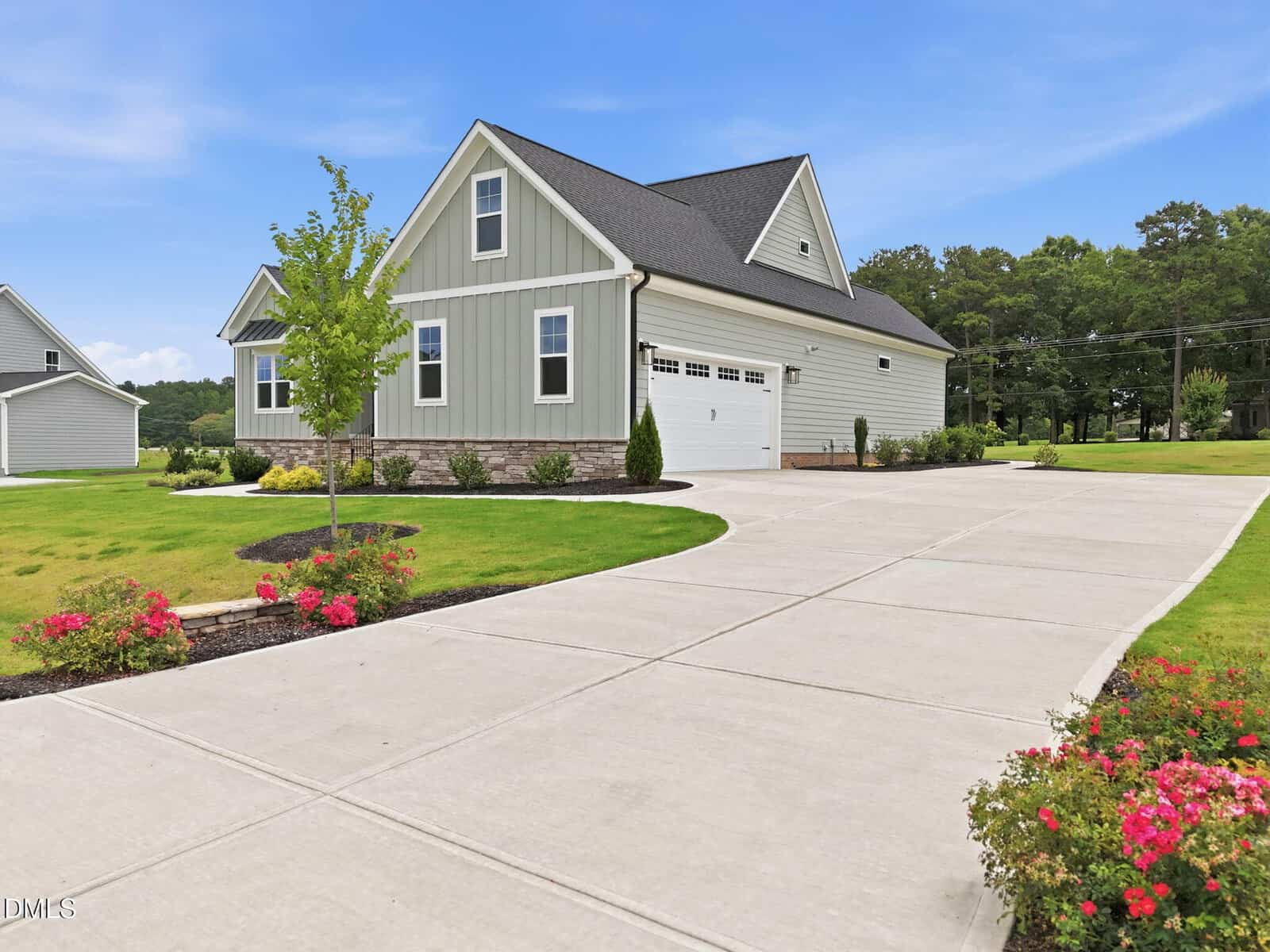 A modern gray house with white trim, a two-car garage, and a large concrete driveway. The yard features manicured lawns, young trees, small bushes, and colorful flower beds under a clear blue sky.