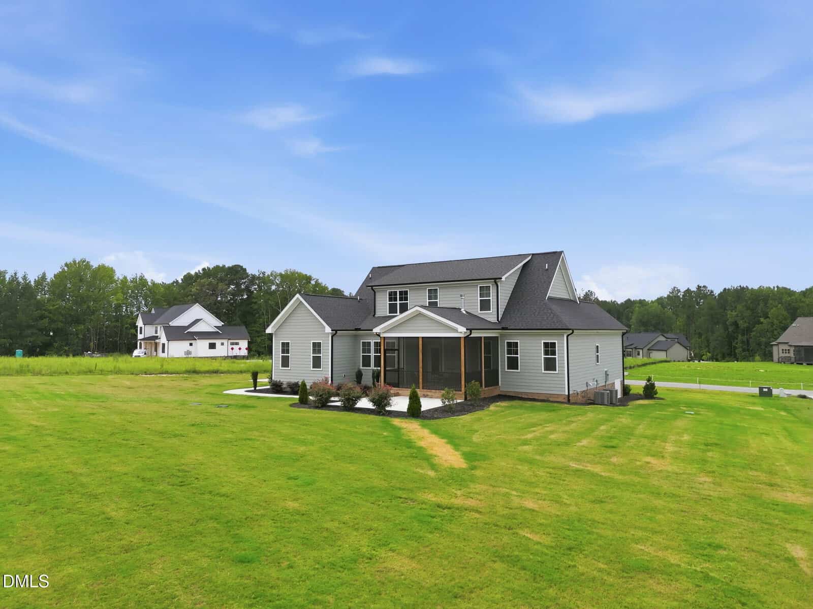 A modern two-story house with gray siding and a screened porch sits on a large, well-maintained lawn with trees and other houses in the background under a blue sky.