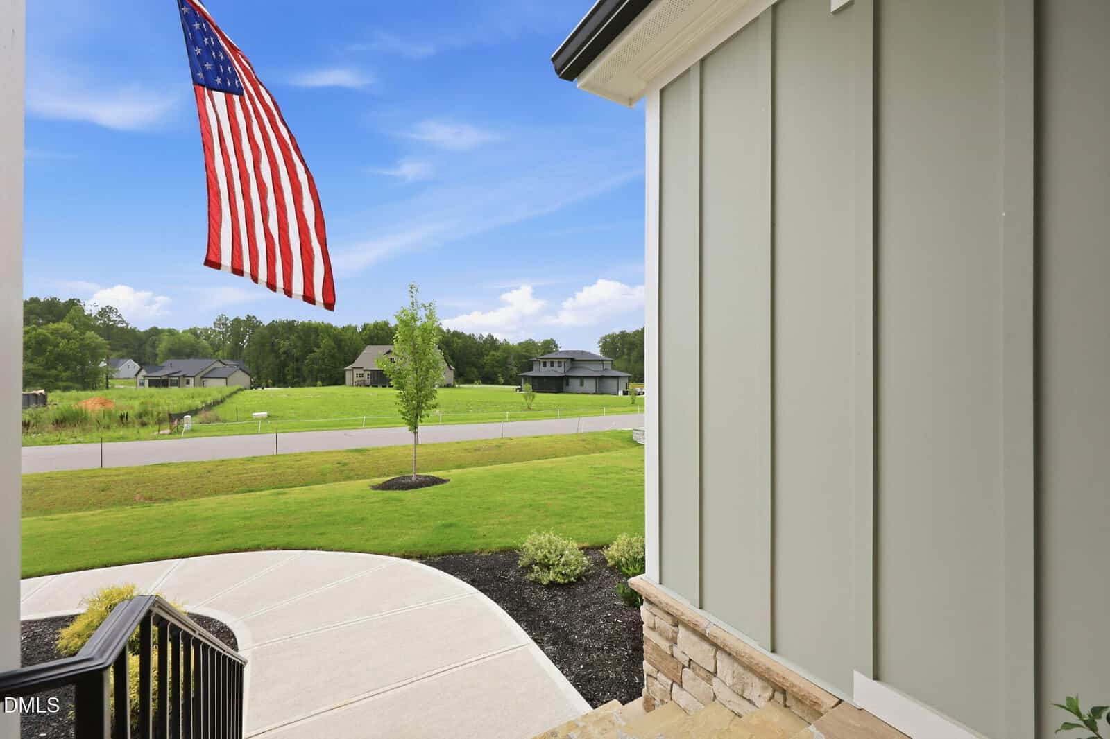 View from a porch with a waving American flag, overlooking a green lawn, sidewalk, road, and several houses in a suburban neighborhood under a bright blue sky.