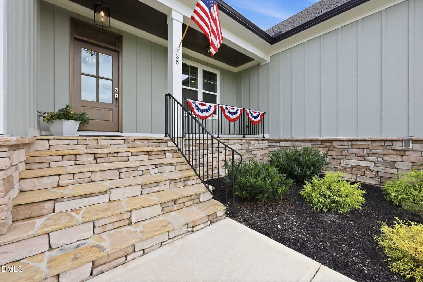 Stone steps lead to a front porch with a brown door, potted plant, and patriotic bunting. The home exterior features light vertical paneling and stonework, with green shrubs planted along the walkway.