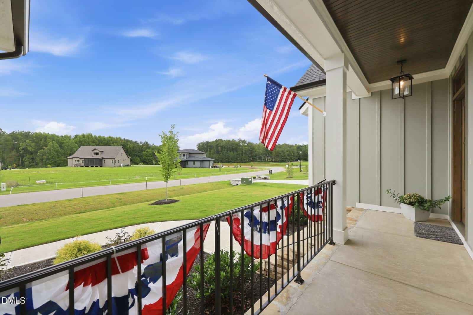 View from a porch decorated with patriotic banners and a U.S. flag, overlooking a green lawn, trees, a road, and houses under a blue sky with scattered clouds.